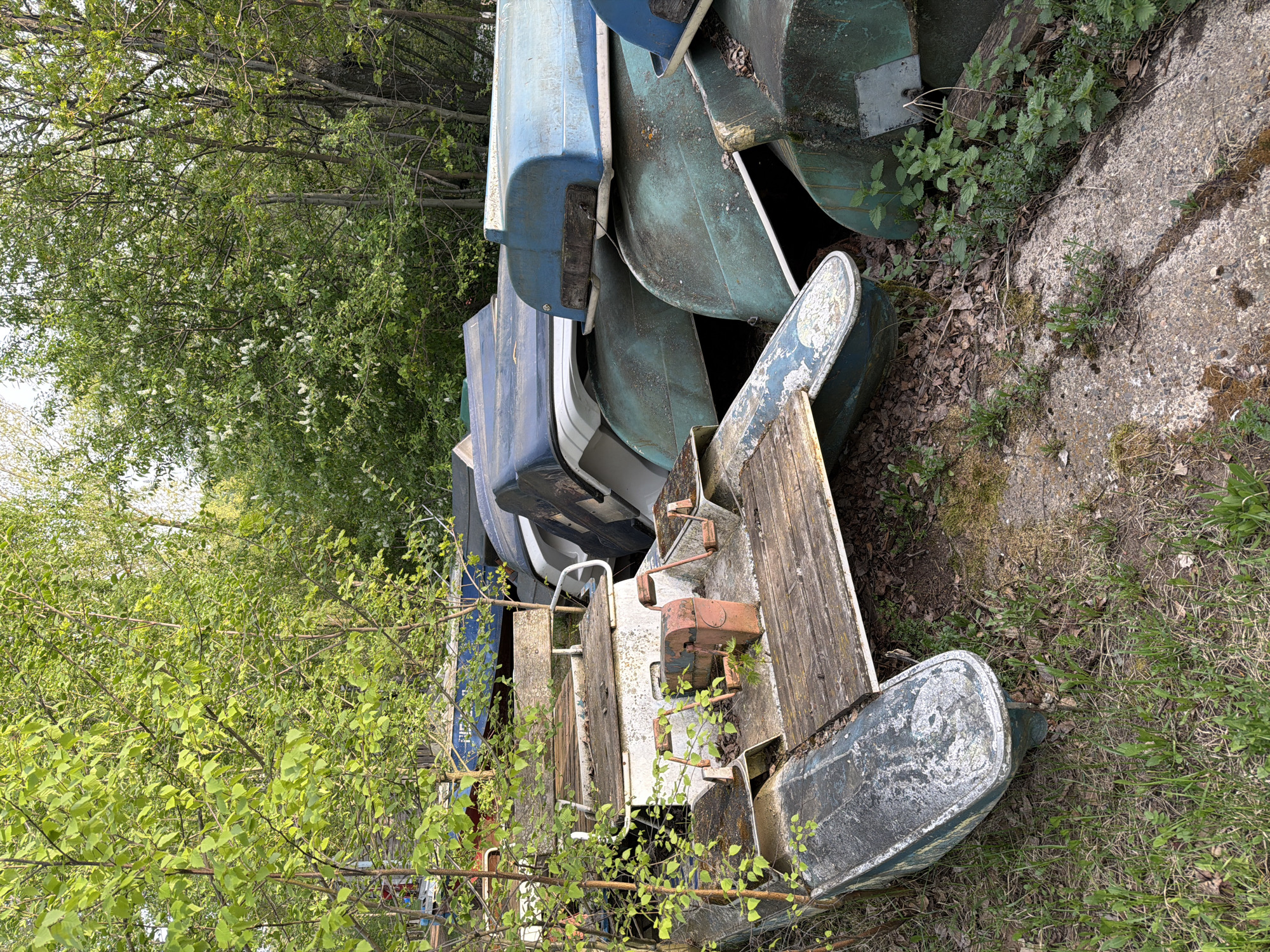 The image shows a pile of old, abandoned paddle boats in a grassy, overgrown area. The boats are stacked haphazardly, with some lying on their sides and others piled on top. Their colors are faded and peeling, suggesting they have been left exposed to the elements for a considerable time. The ground around the boats is covered in grass, weeds, and fallen leaves. The location appears to be a neglected or forgotten area on the outskirts of the city.