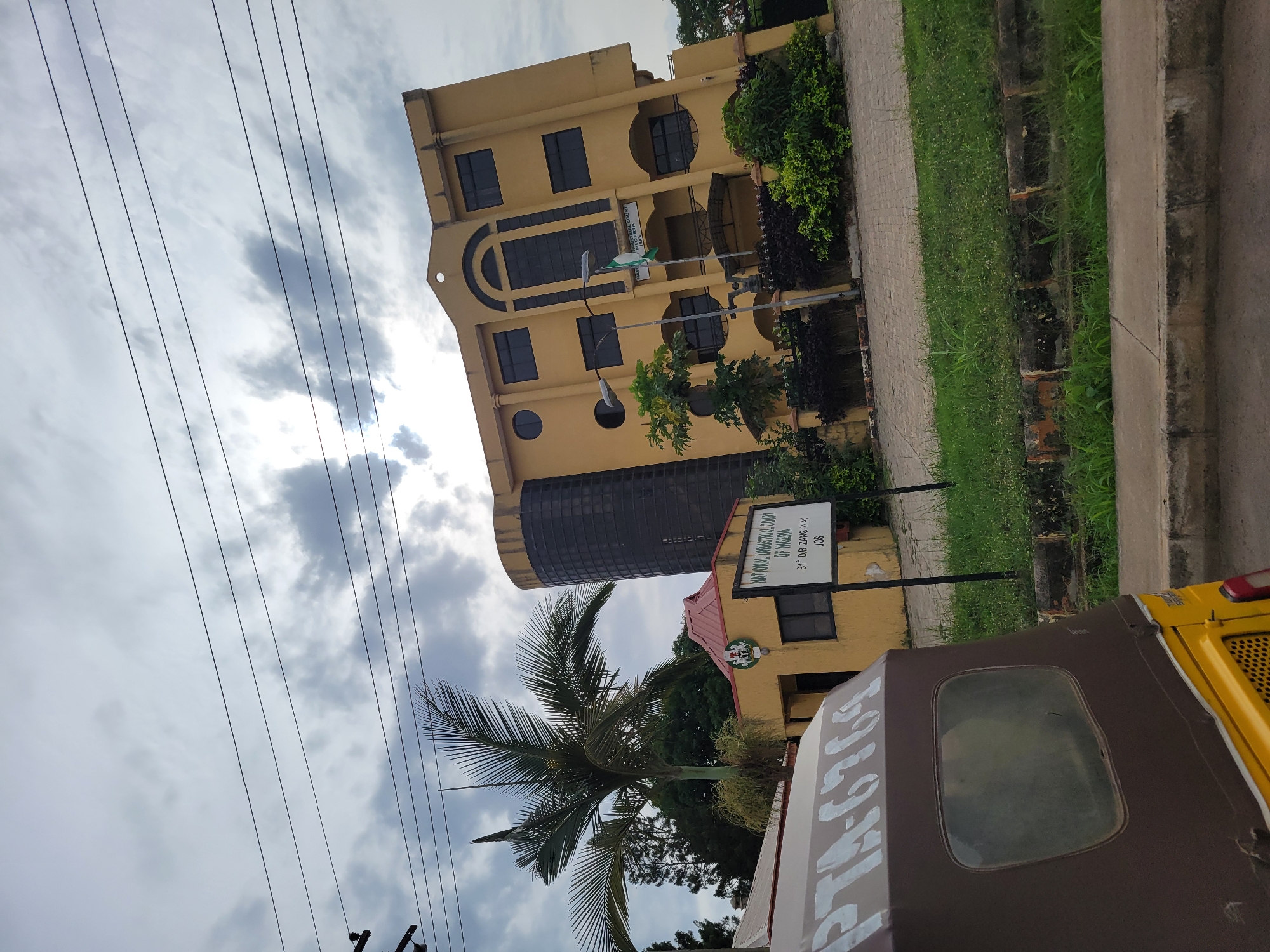 A static street-level photograph captures a yellow, multi-story building under an overcast sky. The building is identified by a sign as the "NATIONAL INDUSTRIAL COURT OF NIGERIA," located at "31st D.B ZANG WAY, JOS." The building features several dark-paned windows, including a large arched window structure on its front facade and some smaller circular windows.
In front of the building, there are various green bushes and a prominent palm tree. A paved sidewalk runs alongside the building, bordered by a strip of grass and a concrete curb. Partially visible in the bottom right corner is a brown and yellow tricycle, likely a keke napep, which has the text "PTA-6769" written on its side. Numerous overhead utility lines stretch across the cloudy sky, which appears bright despite the cloud cover, indicating daytime. The scene depicts a quiet street or institutional area, characteristic of an urban or semi-urban environment in Nigeria, specifically the city of Jos. No human figures or discernible activity are present.