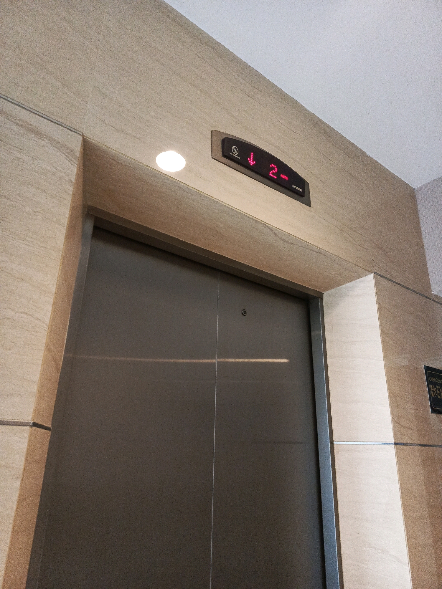 An elevator door is shown closed. Above the door, a digital display shows a downward-pointing arrow and the number "2" in red, indicating the elevator's current floor and direction. The elevator surround and wall are made of light-colored stone tiles. To the right of the elevator, a black sign with white text that reads "EMERGENCY EX" is visible. The setting appears to be an indoor public space, likely a building lobby in Tegal, Indonesia.