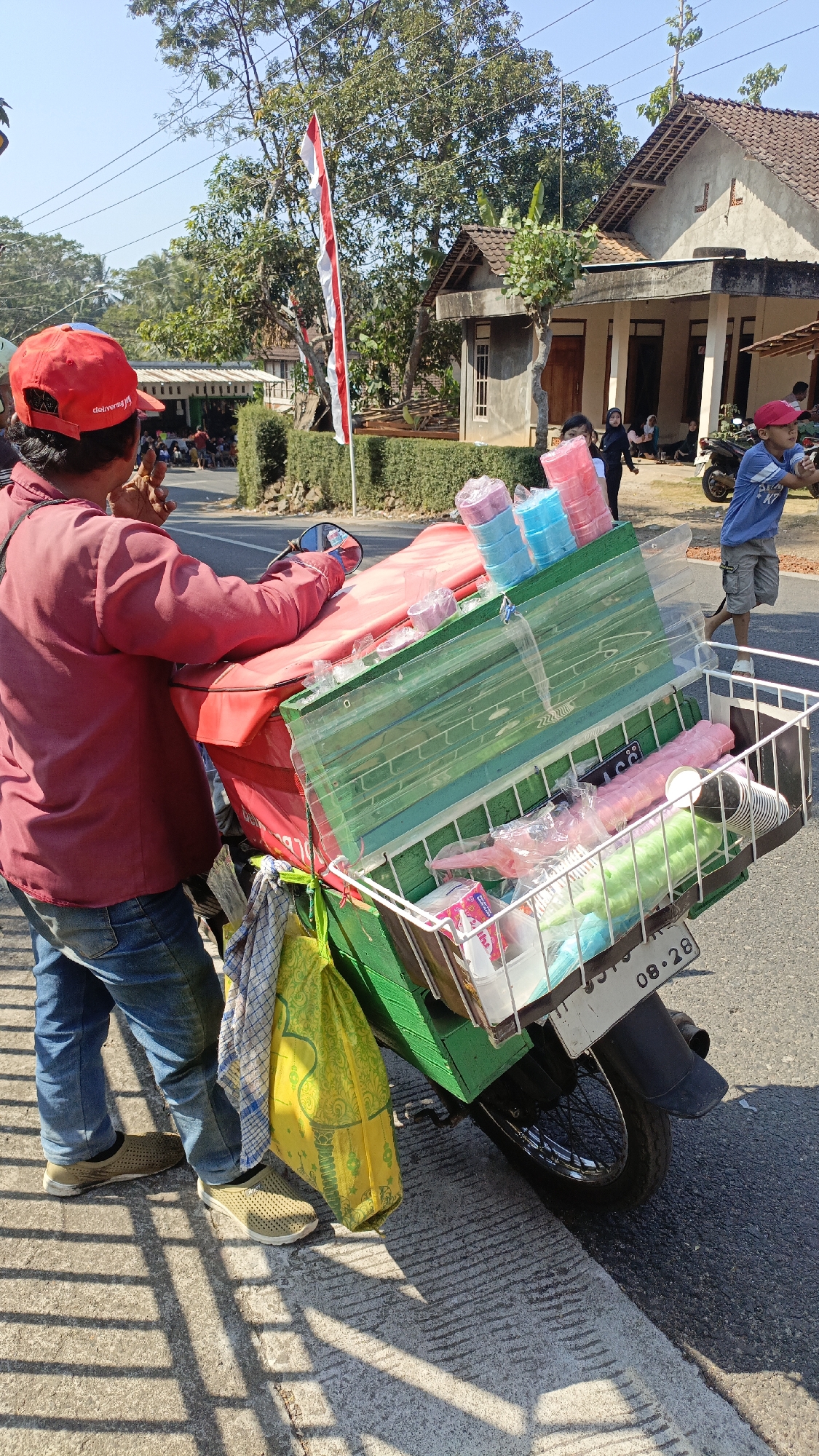 In Krajan Lor, Indonesia, on a sunny day, a person wearing a red cap and a maroon shirt stands next to a motorcycle. The motorcycle is modified with a green cargo box and a white wire basket attached to the rear, carrying various items. These include stacks of colorful plastic cups in blue and pink, plastic wrappers, and a yellow reusable shopping bag. The person appears to be interacting with something off-camera, holding what looks like a small object to their mouth. In the background, a residential building with a tiled roof and a white-painted facade is visible, along with trees and a red and white Indonesian flag on a pole. Other individuals, including a child in a blue shirt and shorts, are present in the scene, suggesting a public area. The ground is paved with concrete and asphalt.