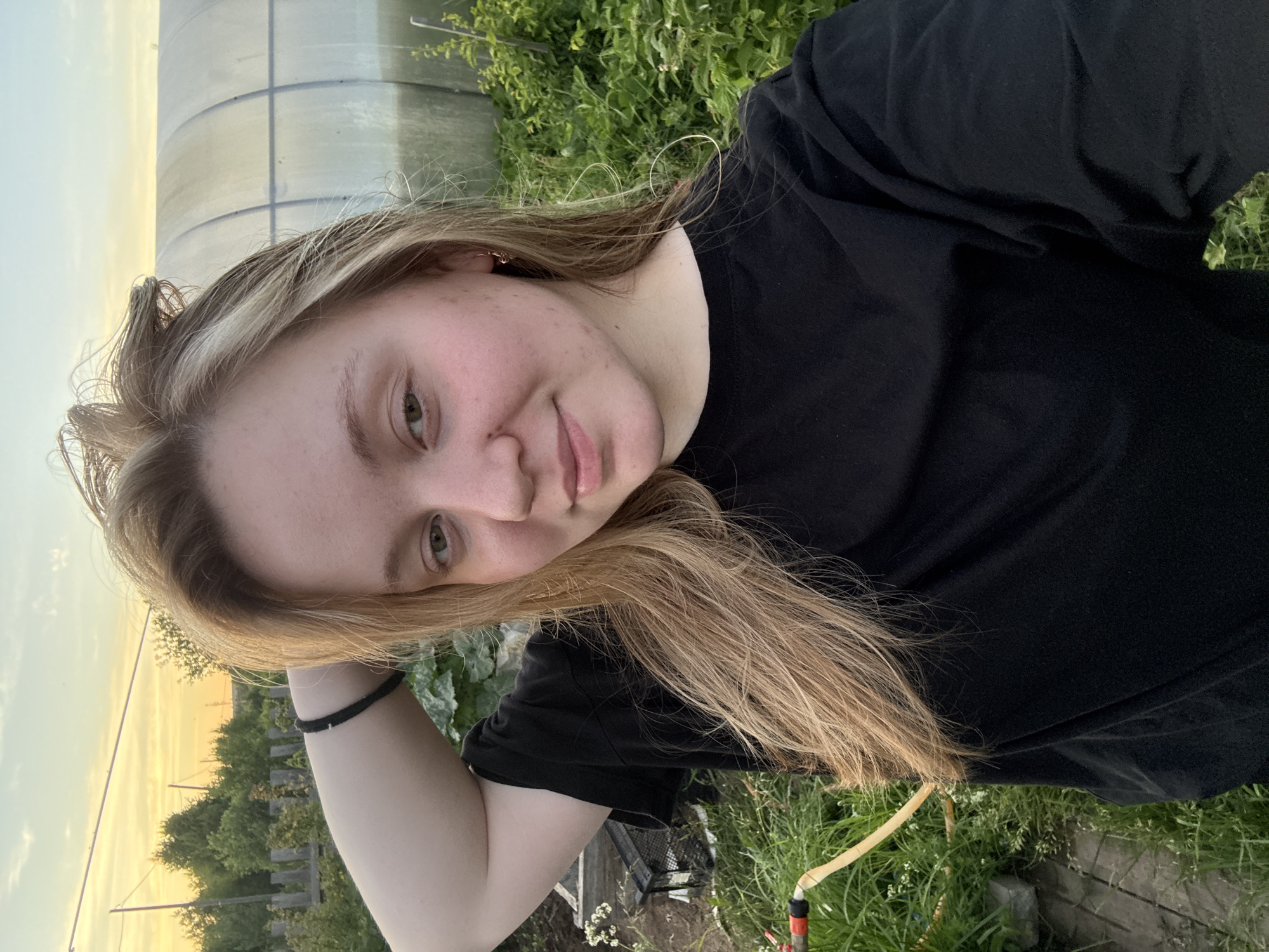 A young woman with blonde hair, wearing a black t-shirt and a black bracelet, is taking a selfie outdoors. Her left arm is raised, with her hand behind her head. She is in what appears to be a garden or rural setting, with lush green vegetation, a greenhouse, and a wooden fence visible in the background. The sky is a soft blend of orange and blue, suggesting it is either sunrise or sunset. A yellow garden hose lies on the ground in the lower left portion of the image. There are no other people or animals present. The location is described as the city of Гаревая, Russia.