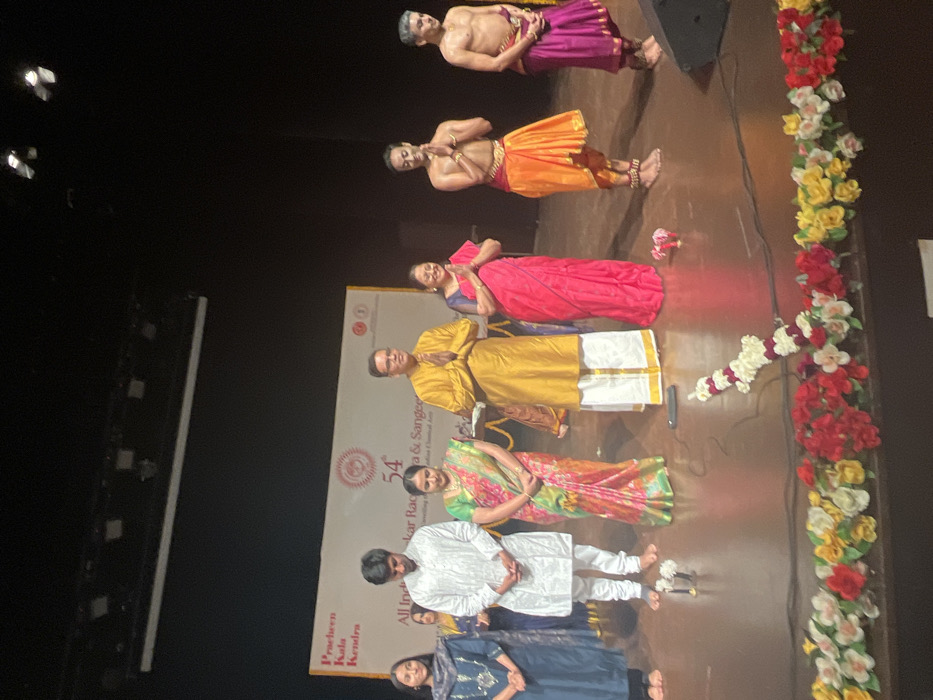 The image shows a stage with seven people standing on it. Six of them are looking towards the front of the stage, while the seventh person is facing the right side of the stage. There is a banner with the number 54 on it. It says "54th All India Festival of Classical Arts 2023". A string of flowers is placed on the stage in the center, and flowers are spread across the front of the stage. The scene is lit by stage lights, and the background is dark. The location is likely the Pracheen Kala Kendra in Sector 35, Chandigarh, India. The coordinates provided (latitude: 30.733, longitude: 76.79) correspond to a location near the center of Chandigarh, within walking distance of the Pracheen Kala Kendra.