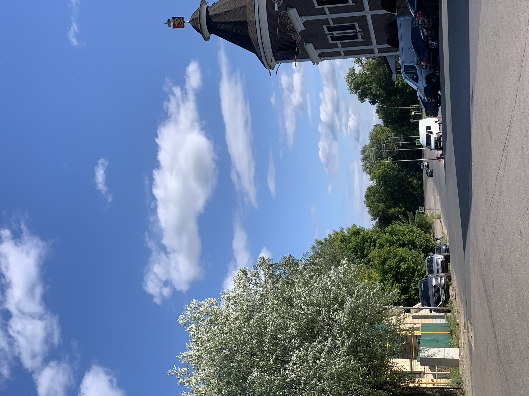 The image shows a street scene in Pavlovo, Russia, on a clear, sunny day. The sky is bright blue with scattered white clouds. In the foreground, a large tree with silvery-green leaves dominates the left side of the frame. Beyond the tree, a building with a dark, conical roof topped with a small, decorative flag is visible. Adjacent to this building, there are several cars parked along the side of an asphalt road, which runs diagonally across the right side of the image.
The parked vehicles include a white van, a dark-colored SUV, and other cars. The road appears to be a relatively quiet street. Green trees line the opposite side of the road, creating a sense of a suburban or semi-rural environment. The building with the conical roof has windows and a facade that suggests it might be a historical or distinctive structure. The overall impression is one of a calm, ordinary day in a Russian town. There is no apparent activity or event taking place, other than the presence of parked vehicles and the natural setting.