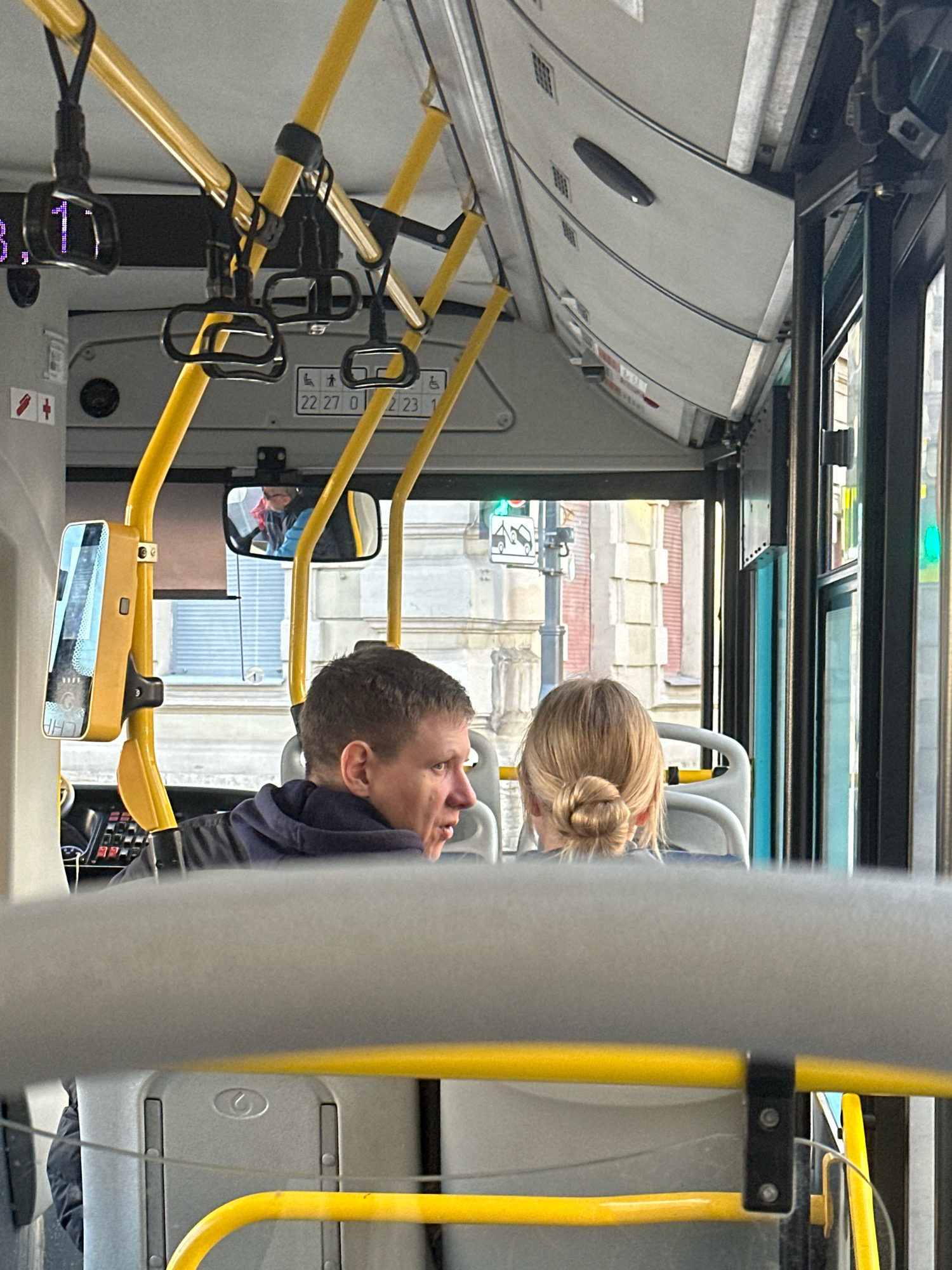 The photograph captures the interior of a public transportation vehicle, possibly a bus, in Saint Petersburg, Russia. The perspective is from a seated passenger looking forward. In the foreground, a section of a gray, upholstered seat back is visible. Immediately behind it, two yellow support poles extend upwards, one to the left and another more centrally.
The primary subjects are two individuals seated further forward. On the left, a man with short, light brown hair is seen in profile, facing towards the right and slightly downwards. He is wearing a dark blue hooded jacket. His mouth is open, suggesting he might be speaking.
To his right, a woman with blonde hair tied in a bun is facing away from the camera. She is wearing a dark top. Both individuals appear to be passengers.
Above and between them, a rearview mirror is positioned, reflecting a person in the driver's area, wearing glasses and a jacket. Yellow handrails and black handles are attached to the poles, characteristic of public transport.
Beyond the passengers, the interior of the vehicle continues with more seating and structural elements. Through the windows, the urban environment of Saint Petersburg is visible. The buildings appear to be made of brick and stone, with architectural styles suggesting an older part of the city. Traffic lights and street signs can also be discerned through the glass, indicating movement and urban activity. The overall lighting suggests an overcast day.