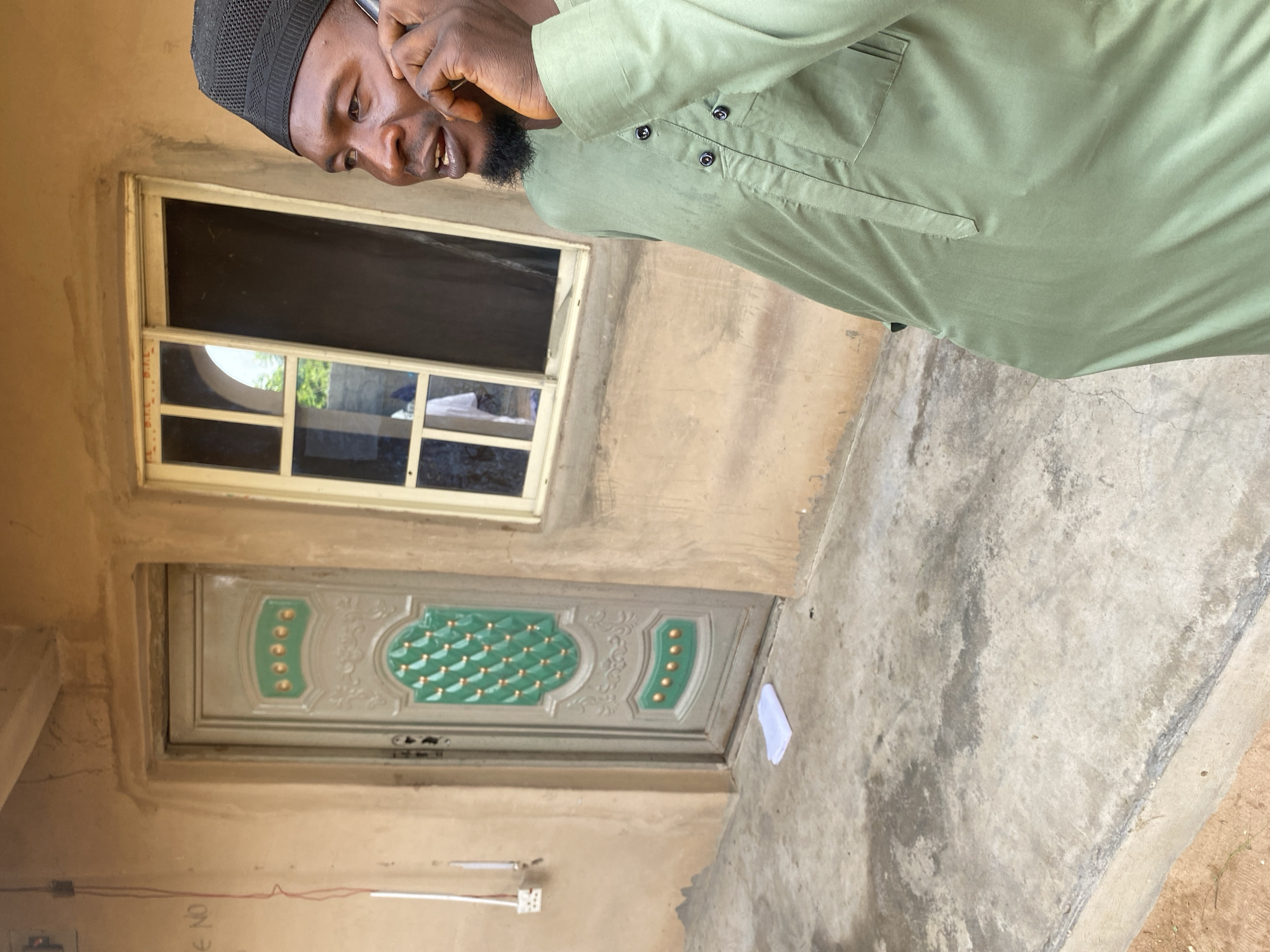 A man in a light green garment and a black cap is standing outside a building and talking on a mobile phone. The building has a tan-colored wall. To the left of the man is a gray door with green decorative elements and gold accents. To the right of the door is a window with multiple panes, one of which is dark. The ground in front of the building is concrete, and there is a folded white object on the ground near the door. The scene appears to be outdoors in a location that could be a residential area.