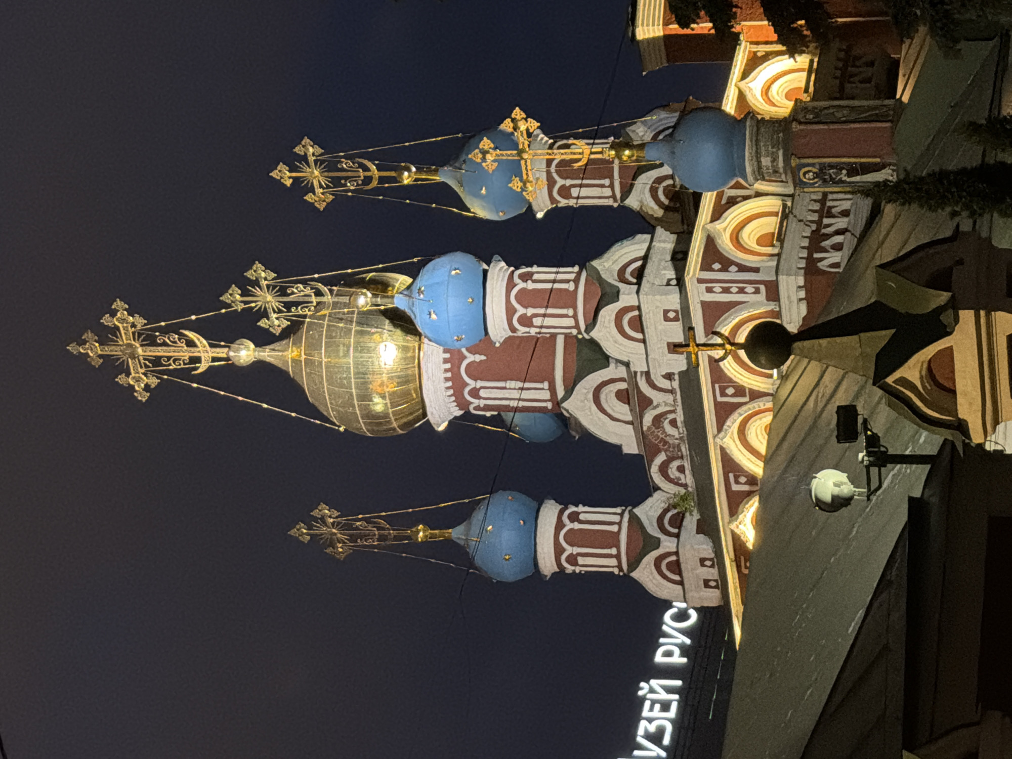 The image depicts the domes of St. Basil's Cathedral in Moscow, Russia, illuminated at night. The domes are painted in vibrant colors of blue, red, white, and gold. The architecture is distinctly Russian Orthodox. Part of the cathedral's exterior wall is visible, showcasing its ornate design. A modern security camera is attached to the wall. In the foreground, there is a neon sign in Russian that reads "МУЗЕЙ РУС", which means "Russian Museum". The background is a dark night sky.