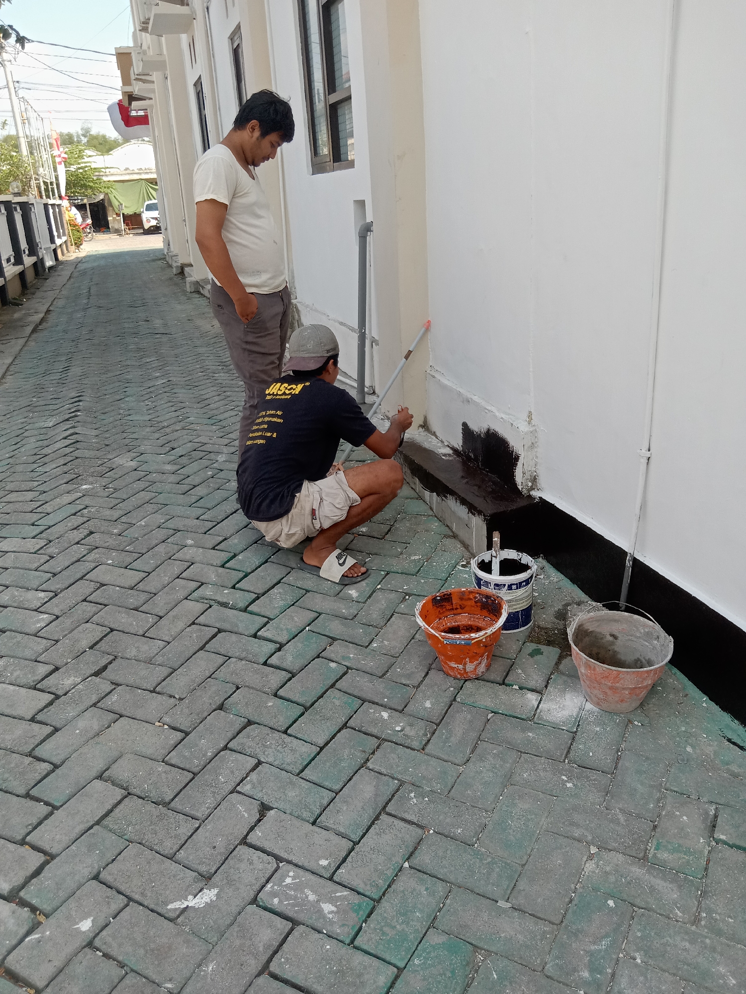 Two men are painting the exterior of a building. One man, wearing a white t-shirt and gray pants, stands with his hands in his pockets, observing. The other man, wearing a black t-shirt, shorts, a cap, and flip-flops, is kneeling and using a paint roller to apply black paint to the base of the building. Three buckets, two of which contain paint, are on the herringbone-patterned paved ground in front of him. The scene takes place outdoors, likely in an alleyway, with a building on the right and a fence and more buildings in the background. The environment appears to be a residential or commercial area in Klepek, Indonesia, suggested by the architecture and vegetation. The lighting indicates it is daytime.
