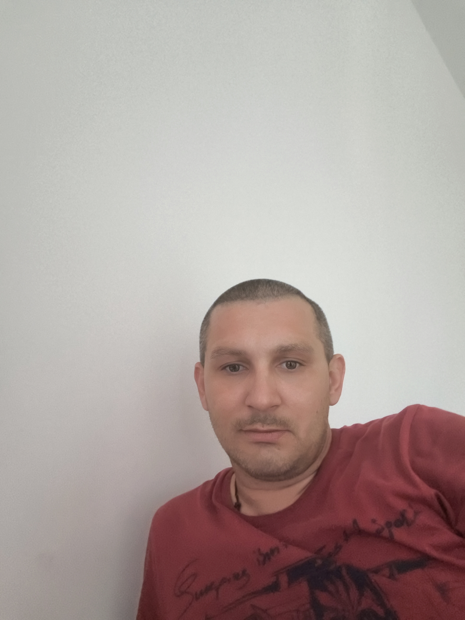 A man with short brown hair and a light complexion is shown from the chest up. He is wearing a red t-shirt with a dark graphic on it. He is looking towards the camera. The background is a plain, light-colored wall. The image appears to be a selfie taken indoors. No specific location in Wolgast, Germany is identifiable.