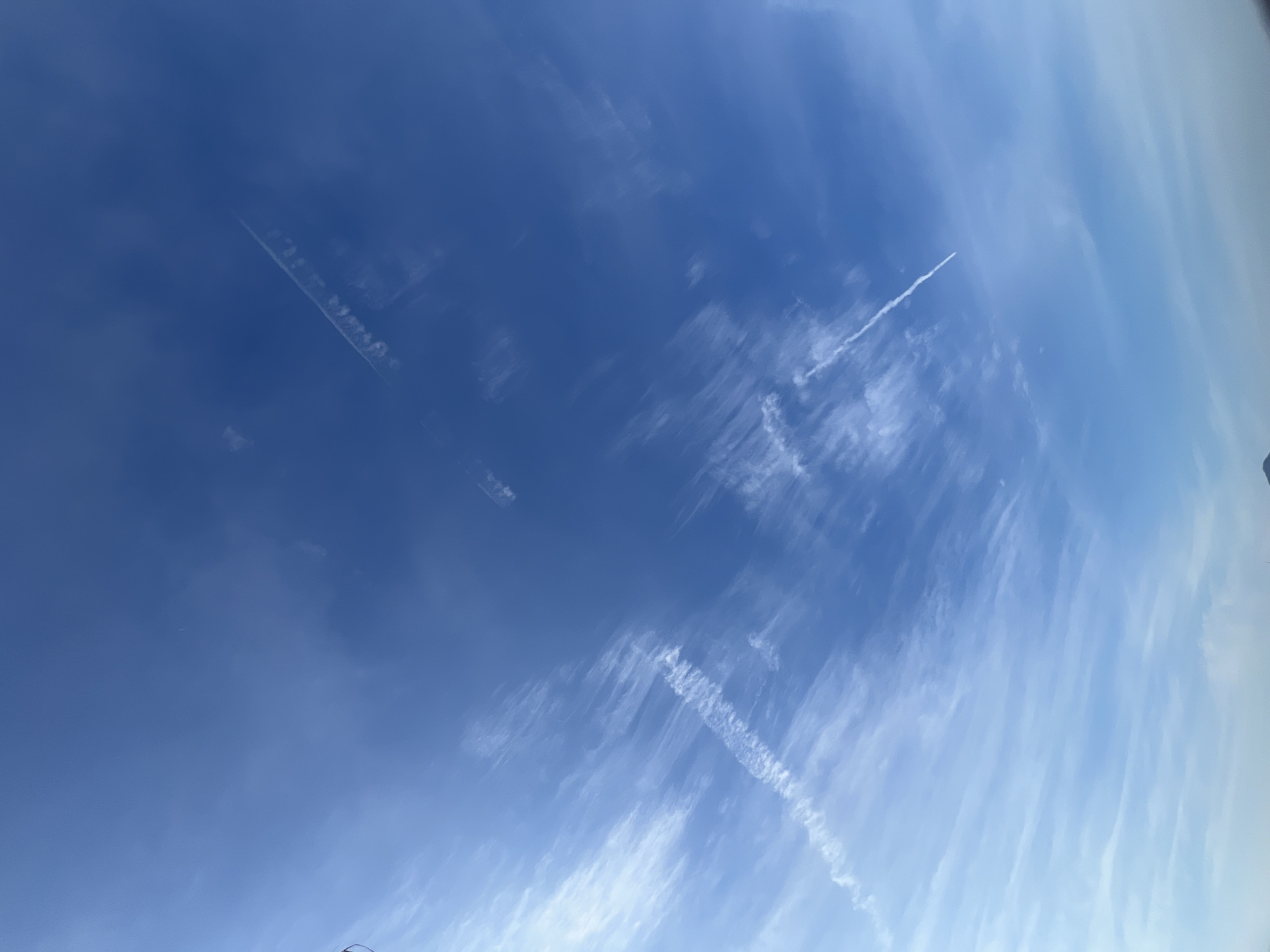 The image captures a clear blue sky above Đà Nẵng, Vietnam, adorned with wispy white clouds and contrails from at least two aircraft. The clouds appear to be cirrus or cirrostratus formations, indicative of high-altitude moisture. The contrails, which are elongated lines of condensed water vapor, suggest that airplanes have recently passed through the upper atmosphere. One contrail is prominent in the lower left quadrant, stretching diagonally upwards. Another is visible in the upper right, also at an angle. The overall scene evokes a sense of openness and altitude. There are no visible people, structures, or ground-level details in the frame.