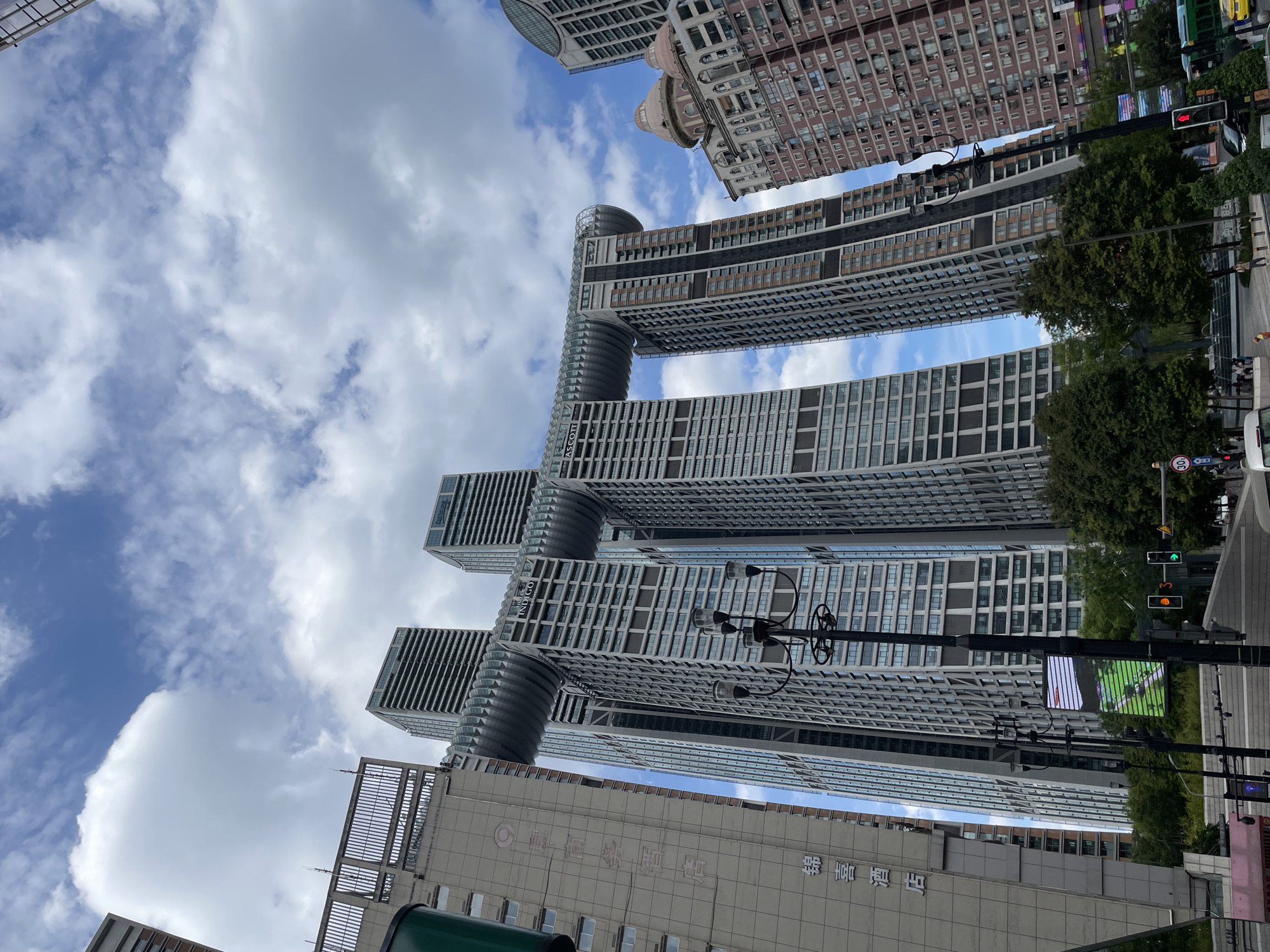 A low-angle, wide shot captures a group of skyscrapers under a partly cloudy sky in Yuzhong District, China. The most prominent structure is a building featuring a massive horizontal connection between its upper sections, resembling a wave or a bridge. This unique architectural element connects at least two, possibly four, tall, modern buildings with glass facades and visible window patterns.
To the left of this central structure, a darker, older-looking building with Chinese characters is partially visible. Further to the right, another building with a red-brick facade and domed rooftops stands out. The foreground is dominated by a street with traffic lights, a street lamp with multiple bulbs, and some trees. The street appears to be an intersection with visible traffic, including a bus and cars, and pedestrian activity on the sidewalk. The sky is filled with dramatic white clouds against a blue backdrop. The scene is captured from a street-level perspective, looking upwards at the imposing architecture.