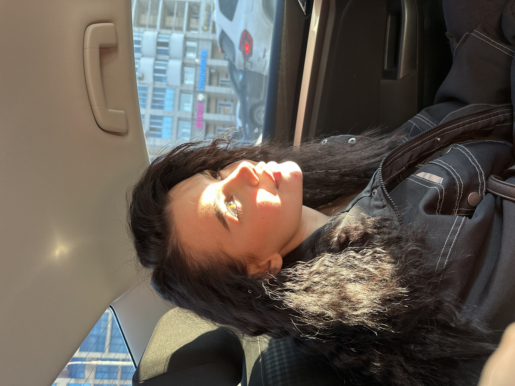 A woman with long, dark hair and light skin is lying in a car. She is wearing a dark jacket with white stitching. The car's interior is beige and black. Outside the car window, there are buildings with blue windows and a sign in Russian that includes the word "KOMANDNYY." Based on the Cyrillic text, the location is likely in Russia. Given the architectural style and information readily available, this is in the city of Kazan, Russia. There is sunlight illuminating the woman's face.