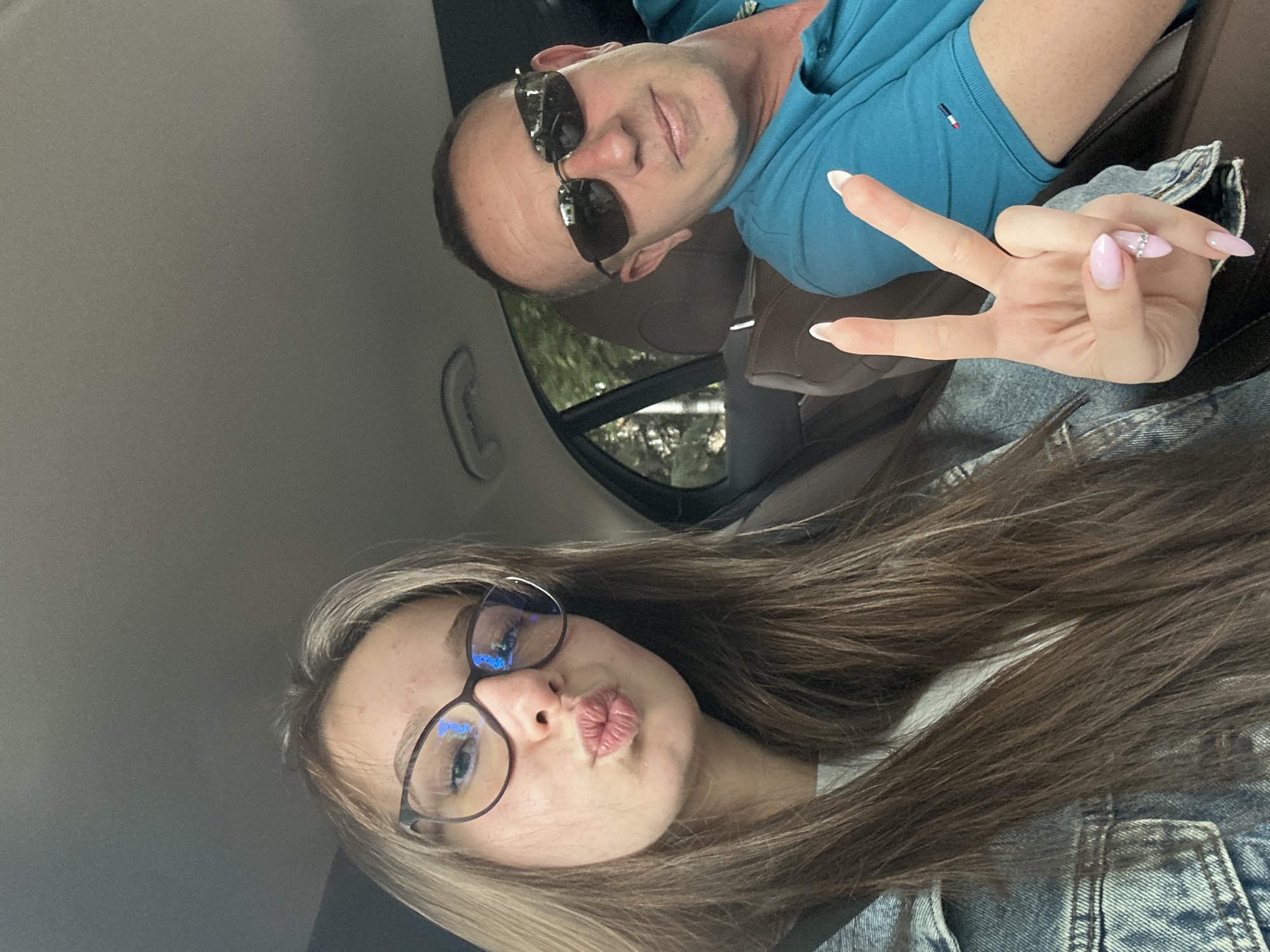 A selfie was taken inside a car by a light-skinned female wearing glasses, a white top, and a denim jacket. Her face is tilted in a kissing expression. The photo also includes a light-skinned male wearing sunglasses and a blue t-shirt, with a hand making a peace sign visible. The location is possibly Blagoevgrad, Bulgaria. The interior of the car is visible, including the headliner, seats, and a glimpse of the outside through the car window.