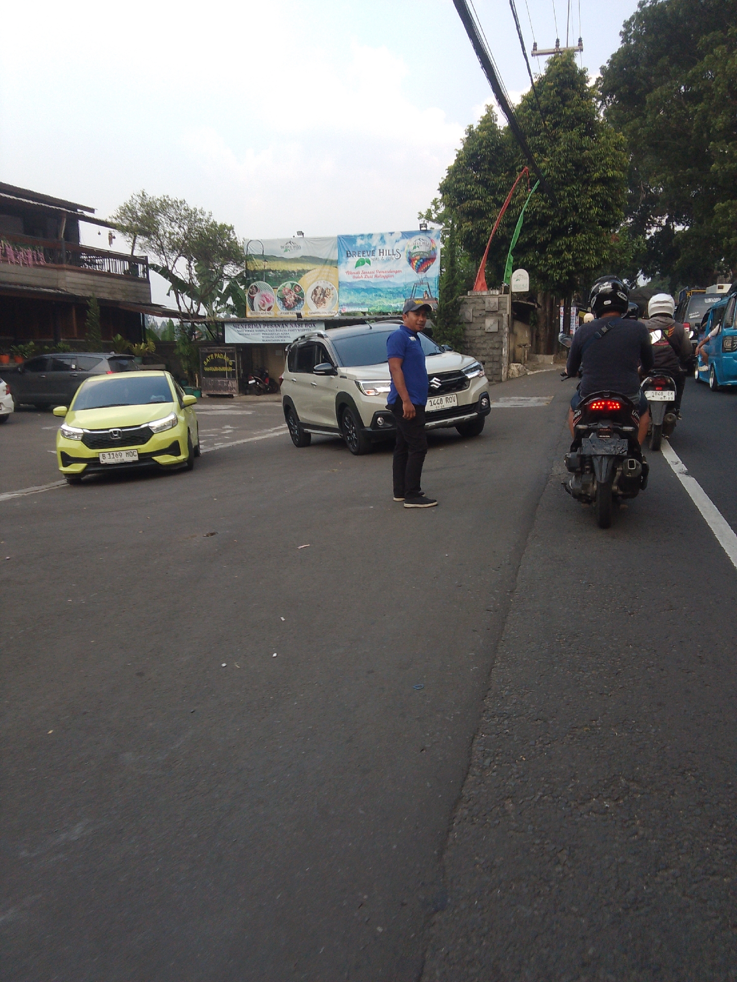 The image depicts a street scene in Cisarua, Indonesia, likely during daytime. The foreground shows a paved road with two lanes marked by a white line on the right. In the middle ground, several vehicles are present. A yellow sedan is parked on the left side of the road, facing forward, with its license plate visible. Behind it, a white SUV is also parked. A man wearing a blue t-shirt and black pants stands next to the white SUV, looking towards the camera.
Further down the road, three motorcycles are visible. The closest motorcycle is facing away from the camera, with its taillights illuminated. Two individuals are riding it, both wearing helmets. Another motorcycle is ahead of it, also with a rider wearing a helmet. To the right of the motorcycles, a blue vehicle, possibly a van or truck, is partially visible.
In the background, there are buildings and lush green trees. A large banner is displayed on a building, featuring text and images related to a location called "BREEZE HILLS". Overhead, power lines are strung between poles. The overall impression is a typical street view in a residential or commercial area of Cisarua.