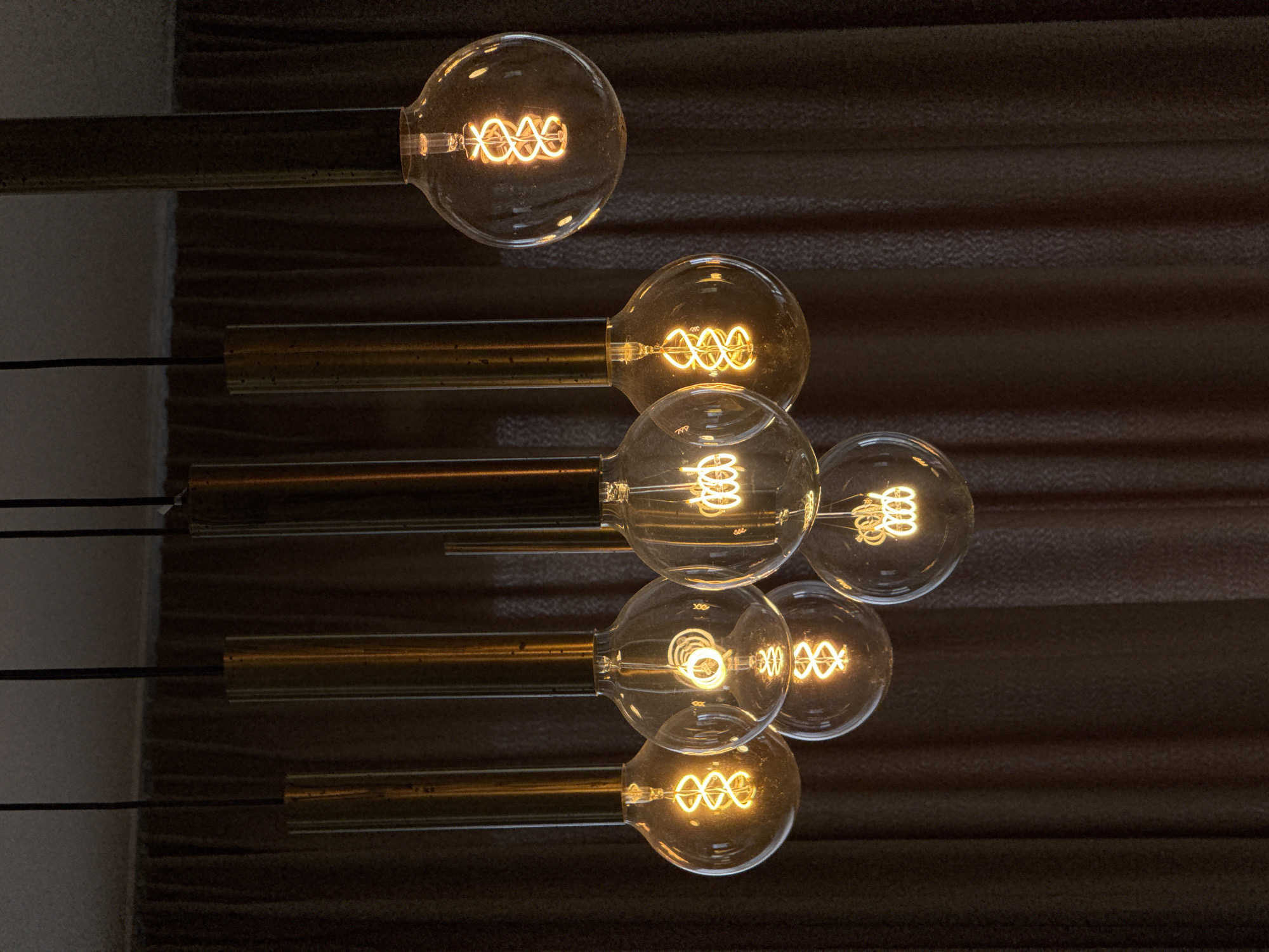 The image shows a cluster of seven lit, vintage-style Edison light bulbs. Each bulb has a clear glass enclosure, revealing a coiled filament glowing with a warm, orange light. The bulbs are attached to horizontal, brushed gold metal rods extending from the left side of the frame. Thin black wires are also visible alongside some of the rods. The background consists of textured, brown panels.
There is no specific information in the image to determine that it is located in the city of Blizikuće, Montenegro.