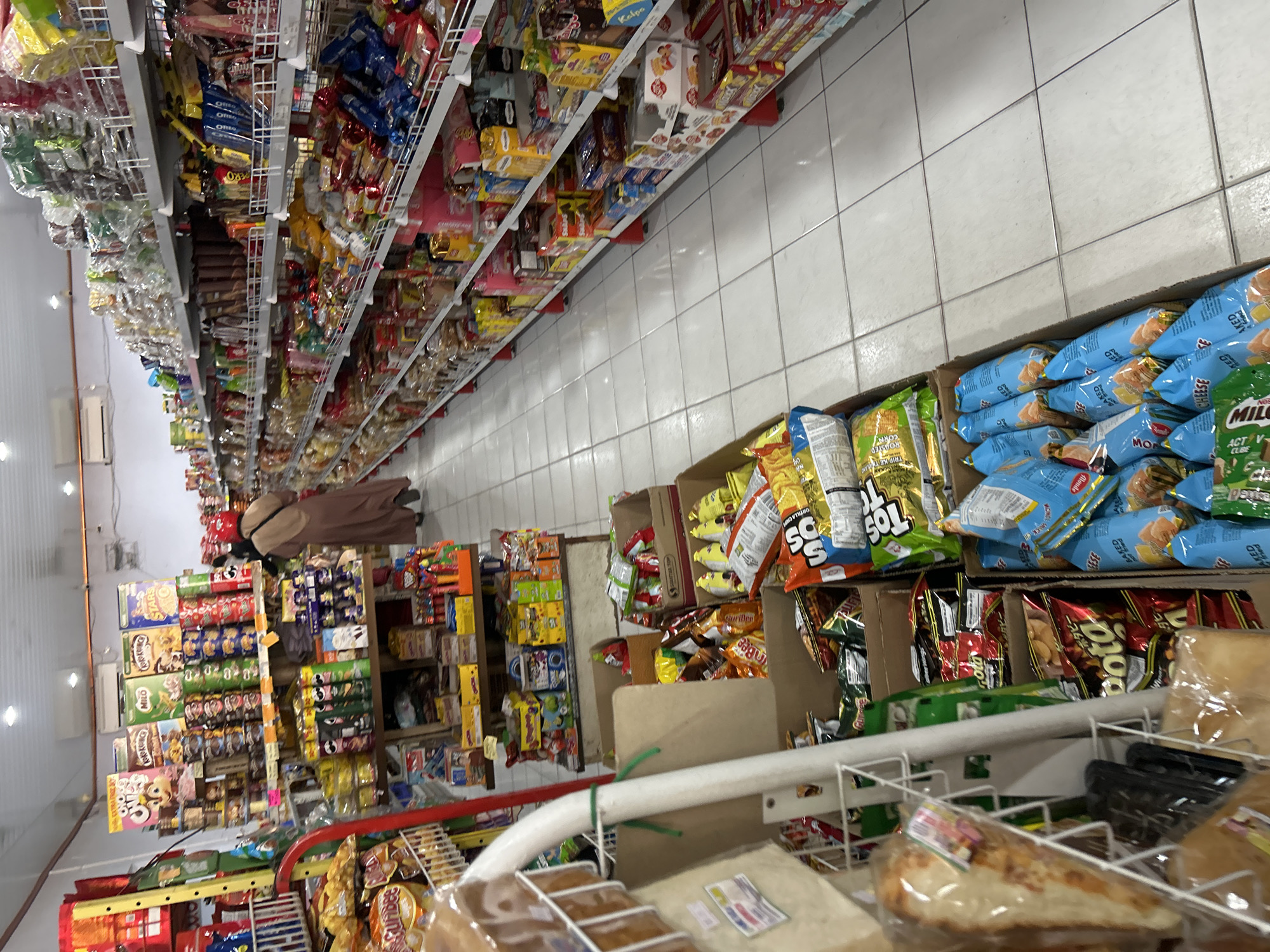 The image shows the interior of a convenience store located in the city of Wonosari, Indonesia. The store features aisles stocked with various packaged food items, including snacks, biscuits, and drinks. In the foreground, several open cardboard boxes are filled with bags of chips and snacks. Further back, shelves are densely packed with a wide assortment of products. A person, partially visible, is walking down one of the aisles. The flooring appears to be tiled. The lighting suggests the store is open for business.