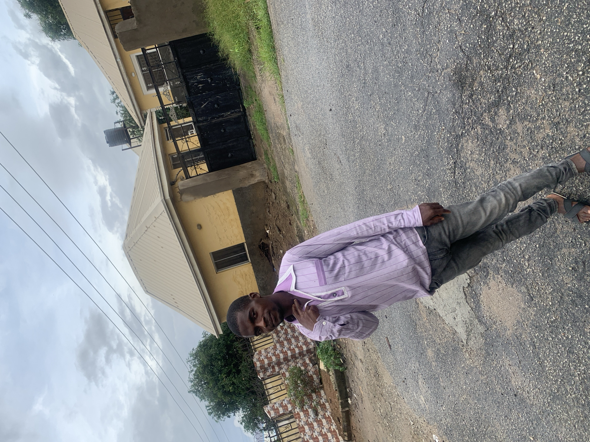 A man in a light purple striped shirt and grey jeans stands on a paved road in front of yellow houses in Abuja, Nigeria. The man is positioned in the center of the frame, facing towards the camera with his left hand raised to his neck. The houses are single-story with beige roofs and black gates. A brick wall is visible to the left of the houses, and trees and grass surround the property. The sky is overcast.