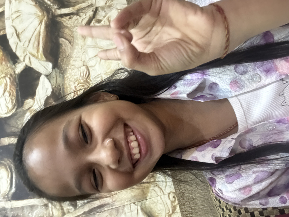 A smiling young woman poses with her head slightly tilted, showing her face clearly. She wears a white shirt with a floral, patterned lavender shirt on top. Her black hair is smooth and shiny. She is wearing a nose ring and earrings, with a bracelet on her wrist. Her hand is up, displaying the peace sign. The background is a mix of textured beige and gold with some patterned fabric.