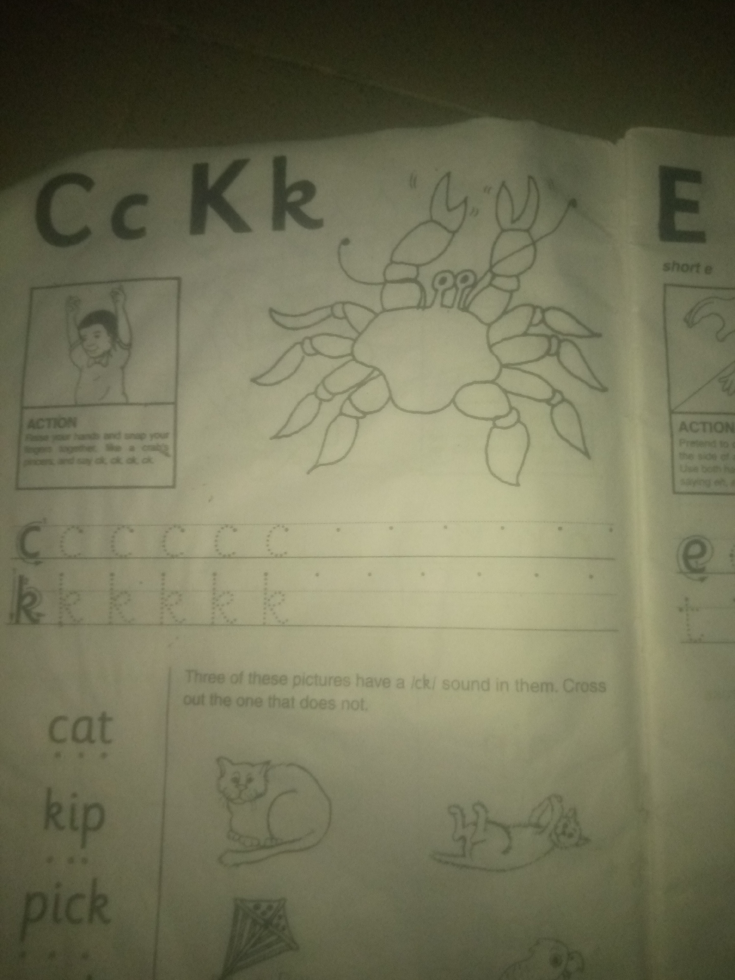 The image is a close-up, angled shot of an open children's workbook page, likely from a learning center in Jauro Yinu, Nigeria, given the educational context. The page features alphabet tracing exercises for the letters "C" and "K," with dotted outlines for children to practice writing them.
To the left of the alphabet section, there's a drawing of a boy with his arms raised, accompanied by text labeled "ACTION" instructing the child to "Raise your hands and snap your fingers together, like a crab's pincers, and say 'ck, ck, ck, ck'." Above this is a larger, stylized drawing of a crab.
To the right, the letter "E" is shown with the text "short e." Below this, there's another "ACTION" instruction which is partially obscured, but mentions pretending to do something and saying "eh, eh, eh." A small drawing of a hand is also visible.
Further down the page, there are words with dotted letters: "cat," "kip," and "pick." Below these words are three illustrations: a cat sitting, a kite, and a cat lying on its back. A text box above these illustrations reads, "Three of these pictures have a /ck/ sound in them. Cross out the one that does not."
The overall environment appears to be indoors, with the lighting suggesting it might be a dimly lit room or that the photo was taken in low light conditions. The workbook page is laid flat. There are no people actively engaged in the activities shown in the image, only static representations. The page itself appears to be made of standard paper.