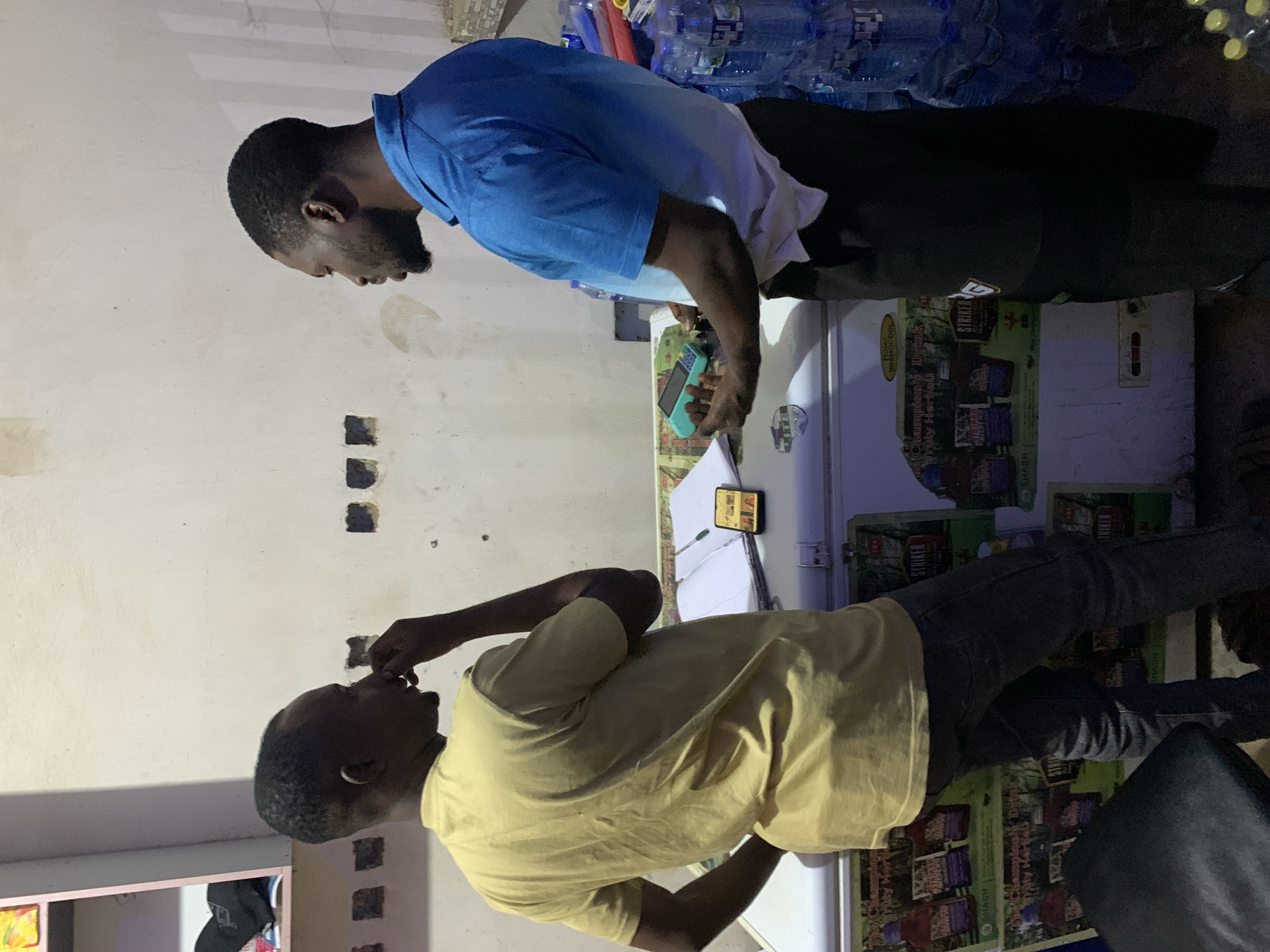 Two African males stand facing each other next to a refrigerator, with one male in a blue shirt using a smartphone. The location is New Stadium, Nigeria.
The male on the left wears a yellow short-sleeved shirt and dark-colored pants. He appears to be looking at his phone on the refrigerator. The male on the right wears a blue short-sleeved shirt, white undershirt, and dark-colored pants. He is also looking at a smartphone. The fridge is stocked with soda beverages. Bottles of water are seen at the top of the fridge. The wall behind them has electrical conduits sticking out.