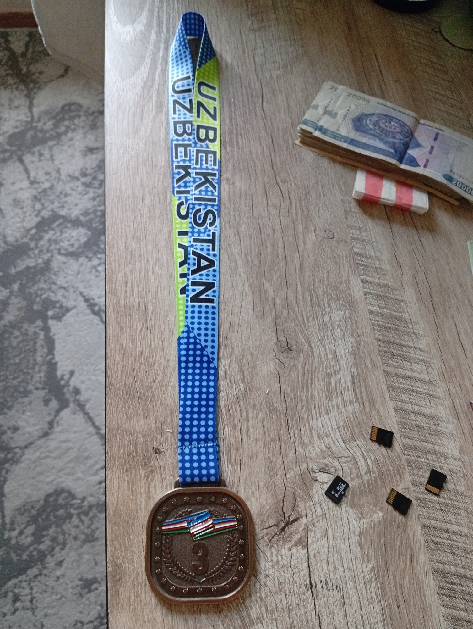 The image shows a medal with a lanyard that reads "UZBEKISTAN" and is adorned with a blue polka dot pattern. The medal itself is bronze colored and features a laurel wreath design with the number "3" prominently displayed in the center. On top of the number "3", there are three small flags. To the right of the medal, there is a stack of Uzbekistani banknotes, appearing to be 20,000 Som denominations, held together by a red and white band. Scattered around the banknotes are four microSD cards. The objects are placed on a wooden surface with a visible grain pattern. The background on the left appears to be a grey and white patterned rug or carpet. There are no people visible in the image. The setting appears to be a domestic or personal space, likely indoors, given the wooden table and carpet. The provided information about the city of Angren, Uzbekistan is not visually inferable from the image itself, as there are no landmarks or specific contextual clues to indicate this location.