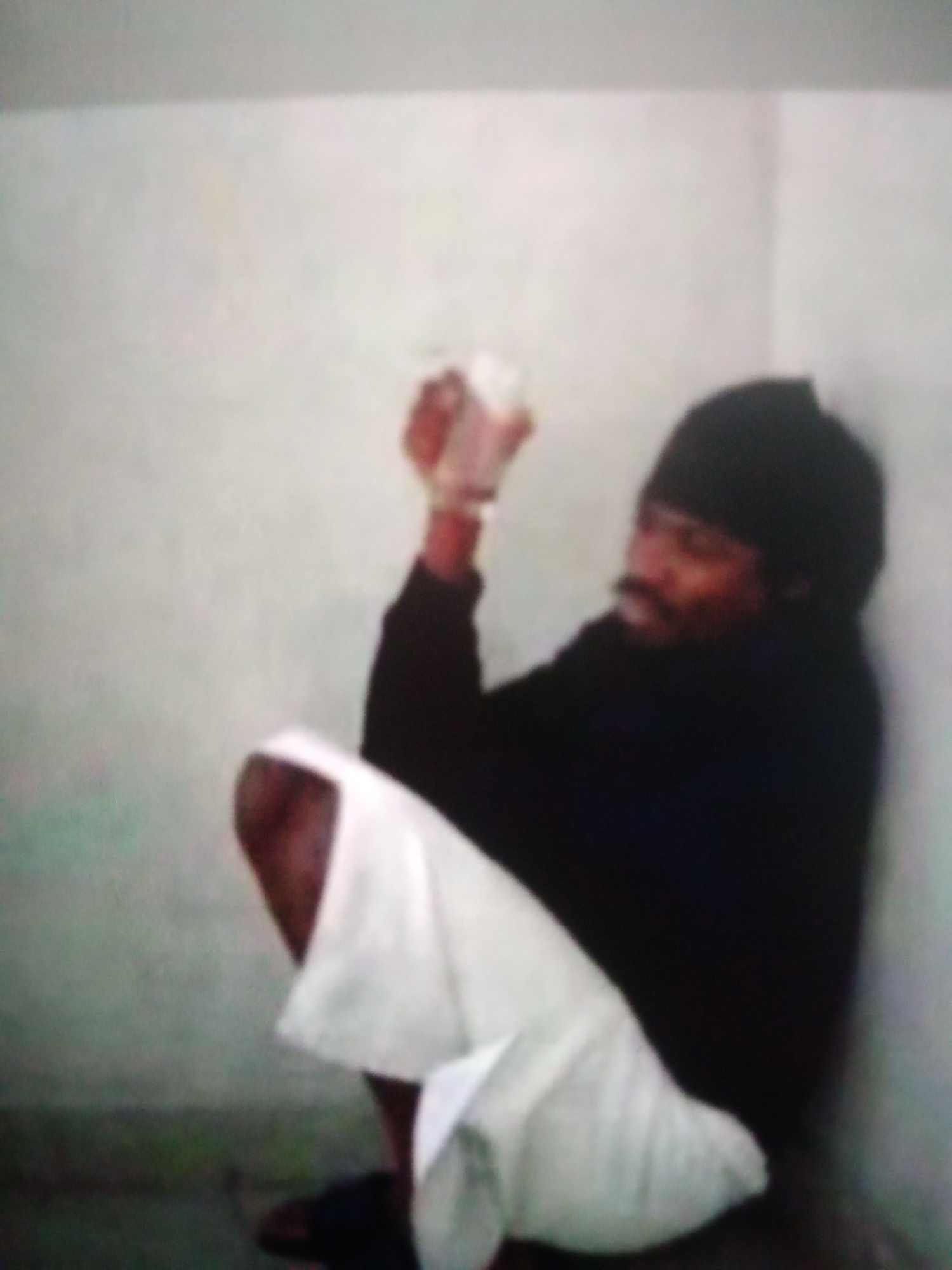 The image depicts a person sitting in a corner against a light-colored wall. The individual is wearing a dark beanie and a black top, with a white garment, possibly a lungi or sarong, wrapped around their lower body. Their knees are drawn up, and they are holding something small and white in their hands, near their face. The person appears to have a mustache and a serious or introspective expression. The scene suggests an indoor setting, possibly a room or a sheltered area, with a plain, undecorated wall and a darker floor. The lighting is somewhat dim, creating shadows and a slightly blurry overall image quality. No text is visible in the image. The context provided indicates Jalingo, Nigeria, which could suggest a cultural or environmental setting relevant to the clothing and appearance of the subject. The activity appears to be a moment of quiet contemplation or engagement with the object in hand.