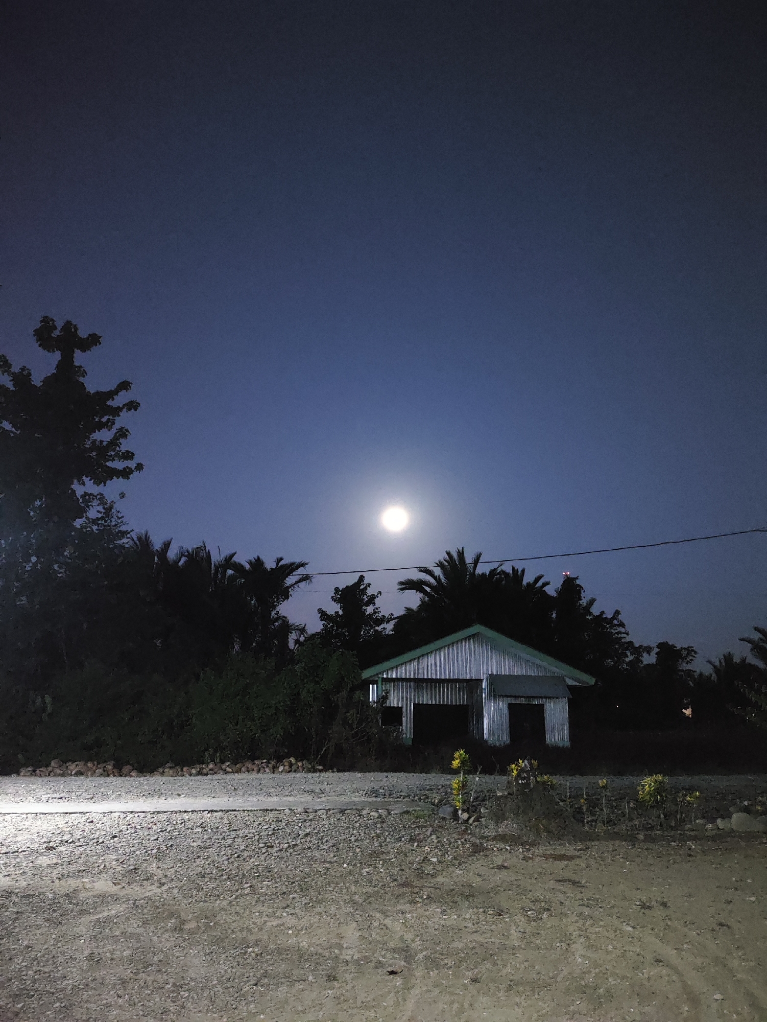 The image depicts a rural scene at night, likely in Jakarta Baru, Indonesia. A full moon shines brightly in a dark blue sky, casting an ethereal glow over the landscape. In the foreground, a gravel road stretches across the frame, illuminated by an unseen light source. To the right of the road, a small, corrugated metal shed stands partially obscured by dark trees and vegetation. The shed appears to be unoccupied and shows signs of wear and tear. The overall atmosphere is one of quiet solitude and natural beauty, with the moon serving as the primary source of light.