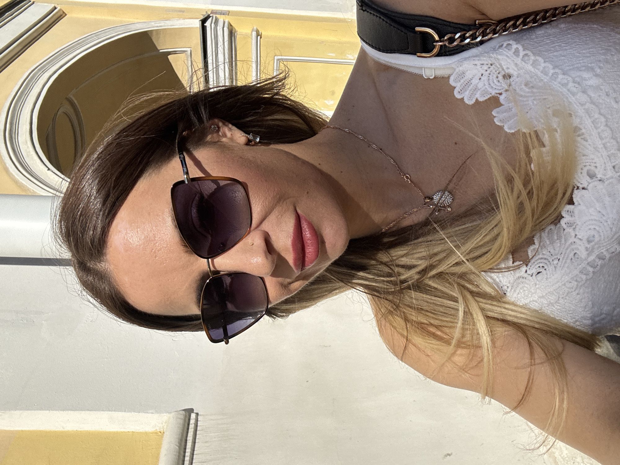 A woman stands outside a building in Saint Petersburg, Russia, on a sunny day. She has long, blonde hair and is wearing large, brown-tinted sunglasses and a white lace top. A gold necklace with a circular pendant is visible around her neck. She is also wearing a small earring in her left ear. The building behind her has a yellow facade with white architectural details, including a large arched opening. The light suggests it is daytime.