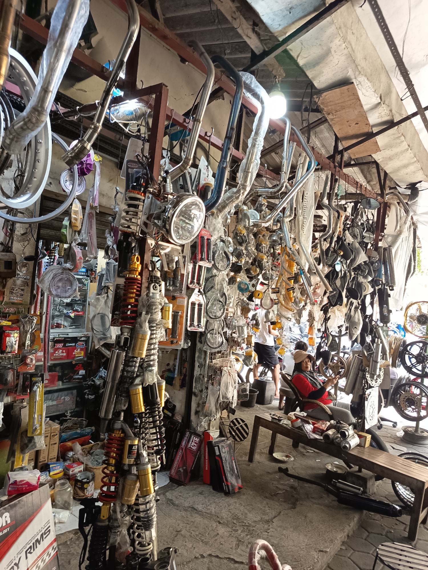 The image depicts an outdoor motorcycle parts stall in Surakarta, Indonesia, characterized by a dense arrangement of various motorcycle components. The stall is filled with an abundance of items, including exhaust pipes, shock absorbers, rims, headlights, and other mechanical parts, many of which are suspended from the ceiling or displayed on racks. The environment appears somewhat cluttered and shows signs of wear and tear, with a rough concrete floor and a corrugated metal roof overhead.
In the foreground, a woman wearing a red shirt and a beige headscarf is seated on a wooden bench, holding a steering wheel. She is looking towards the right side of the frame. Behind her, a person in a white t-shirt and black shorts stands near a motorcycle. The stall is well-lit by natural light filtering through the open structure. The overall scene conveys a busy marketplace atmosphere, typical of a local business catering to motorcycle repair and customization needs in Surakarta.