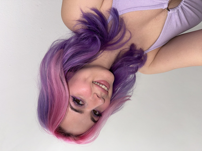 An indoor, close-up photograph depicts a young Caucasian female with prominent, multi-toned dyed hair featuring vibrant purple and bright pink sections. Her hair is styled with waves and falls over her shoulders. She is smiling, revealing upper teeth, with visible dimples on her cheeks. Her eyes are open, looking towards the right side of the frame, and she wears visible eye makeup, including purple winged eyeliner. She is wearing a light purple, ribbed tank top with a plunging V-neckline, secured by a silver-colored O-ring detail at the center of the chest. Her right shoulder is partially visible. The background consists of a plain, light-colored, textured wall. The scene is located in the City of Zagreb, Croatia.