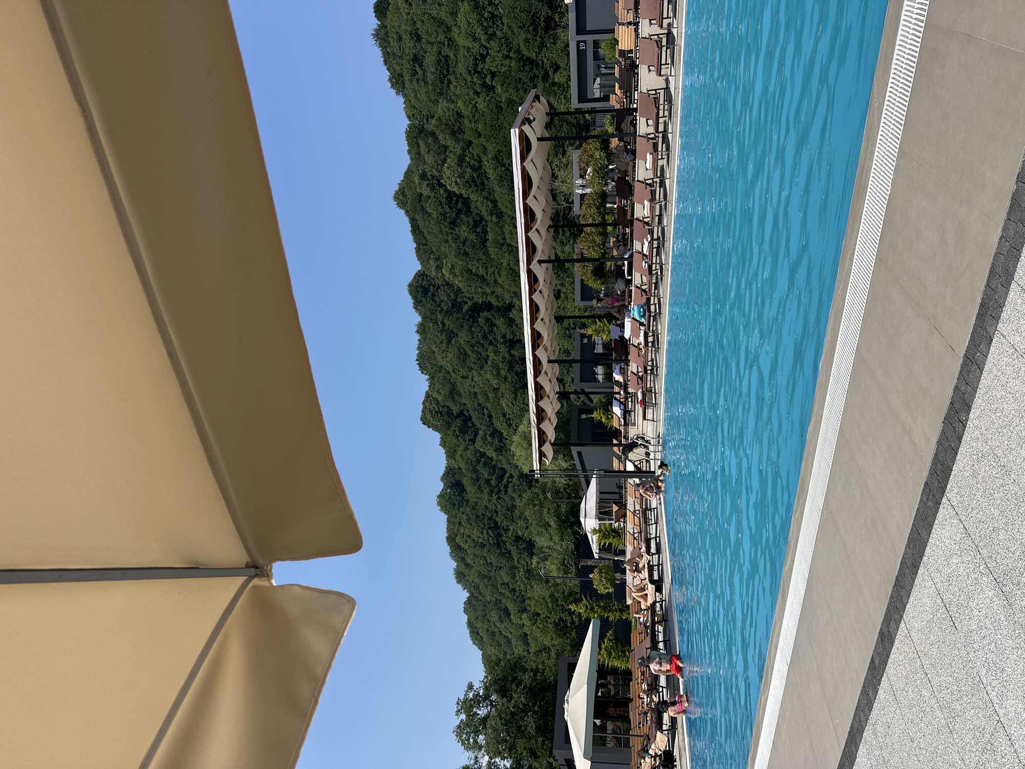 An exterior shot captures a sun-drenched day at a resort, with a bright blue swimming pool dominating the foreground. The pool is surrounded by numerous empty lounge chairs, some with towels neatly placed on them, suggesting a leisurely atmosphere. In the distance, lush green trees cover rolling hills under a clear blue sky, hinting at a natural, serene location. Several people are visible in and around the pool, with some relaxing on lounge chairs and others in the water. A large, light-colored umbrella shades a portion of the foreground, adding to the sense of a comfortable outdoor setting. The scene appears to be a tranquil retreat, possibly in the city of Verkhnyaya Saniba, Russia, known for its picturesque natural beauty.