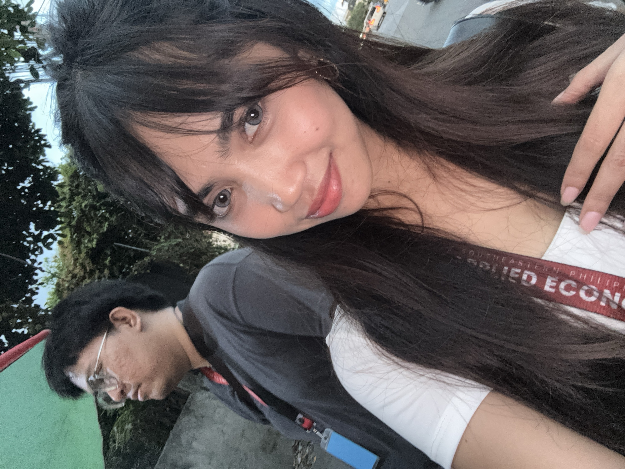 This image is a selfie taken in Davao City, Philippines, with another person in the background. The person in the foreground is a young woman with long, dark hair and light-colored eyes. She is wearing a white t-shirt and a red strap with text that appears to say "SOUTHEASTERN PHILIPPINE". She is holding her hand up near her face. In the background, a person with dark hair and glasses is wearing a dark t-shirt. There is a green tarp and trees visible, suggesting an outdoor setting, possibly near a street given the presence of vehicles in the distance.