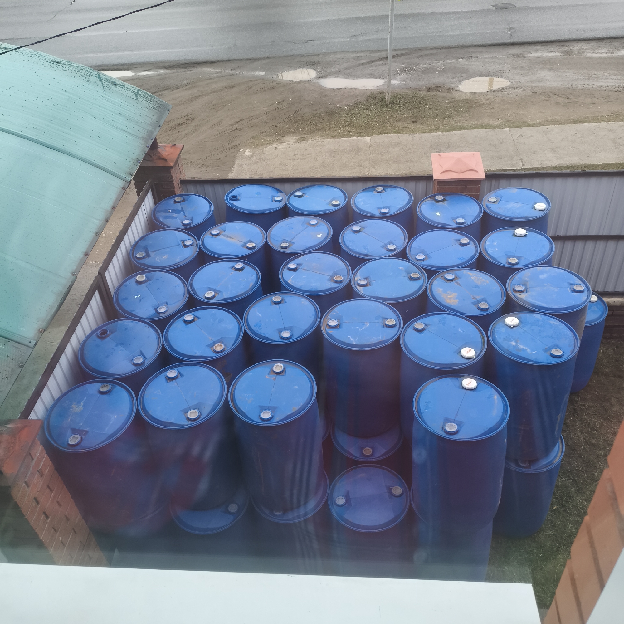 This image, taken from an elevated perspective in Chaikovsky, Russia, showcases a dense arrangement of blue plastic barrels stored outdoors within a fenced area. Approximately 40 to 50 industrial-style drums, characterized by their vibrant blue color and white caps/bungs on their tops, are tightly packed and stacked, some standing vertically and others lying horizontally or at an angle.
To the left, a section of a green, translucent corrugated plastic roof is visible, suggesting a shed or similar structure, with water marks indicating recent precipitation. A brick pillar supports the roof nearby. A metal fence encloses the storage area for the barrels.
Beyond the fence, the ground consists of patches of grass and exposed dirt or mud, featuring several puddles. Further into the background, a gray paved road or street can be seen, also with puddles and tire tracks. A black utility line stretches across the top of the frame.
The scene depicts an overcast daytime, with soft, diffused light and no distinct shadows. There are no people visible, and no discernible activity is taking place; the image captures a static storage setting. No legible text is visible on any of the barrels or surrounding objects.