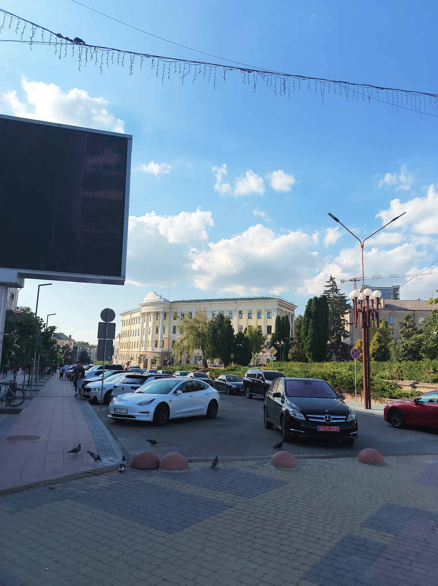 The image depicts a street scene in Ternopil, Ukraine. In the foreground, a paved area with a brick pattern is visible, with several rounded, reddish-brown bollards. Pigeons are present on the ground, walking and pecking. A white Tesla Model 3 is parked in the mid-ground, with a license plate that reads "BO 4994 YA". To the right of the Tesla, a black Mercedes-Benz B-Class is parked, displaying a license plate that reads "03 I C 3271". Further to the right, the rear of a red sports car is visible. In the background, a large, ornate yellow building with classical architectural elements stands prominently. Trees line the sidewalk in front of the building. A large digital billboard is situated on the left side of the image, its dark screen facing upwards. Streetlights and a decorative string of lights are also visible. People are seen walking on the sidewalk on the left side of the image. Construction cranes are visible in the distance above the buildings. The sky is blue with scattered white clouds, and sunlight illuminates the scene.