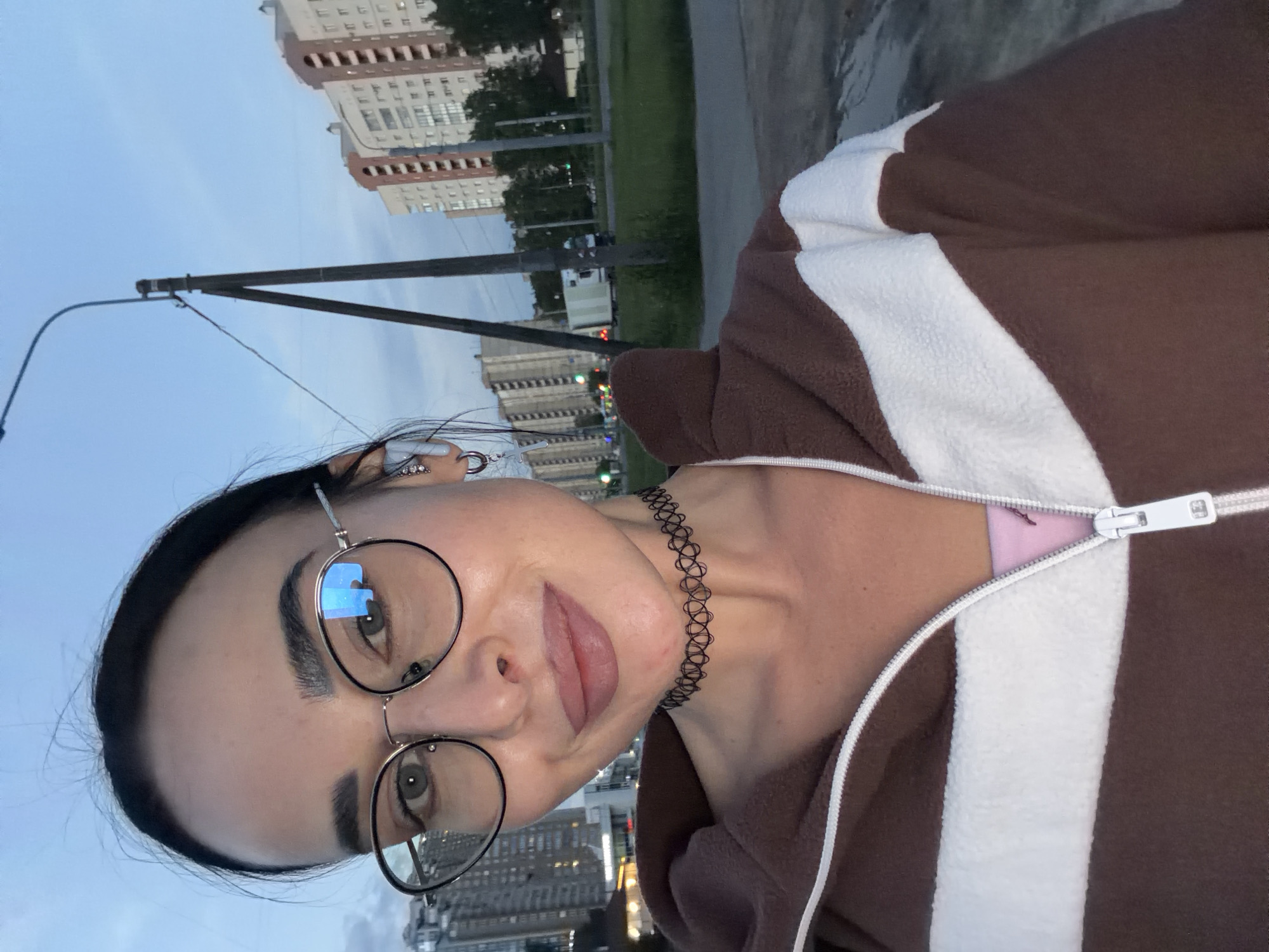 This vertically oriented selfie captures a young woman in an urban setting, likely in Saint Petersburg, Russia. She has dark hair pulled back and is wearing round, metal-framed glasses that reflect a blue light, possibly from a screen. Her expression appears neutral or slightly serene.
She is dressed in a brown fleece jacket with a prominent white stripe running down the front, partially unzipped to reveal a pink garment underneath. A black, patterned choker necklace adorns her neck. Distinctive earrings are visible: one features multiple small jewels, and the other has a circular ring connected to a star-shaped charm by a chain.
The background reveals a city landscape under an overcast sky, suggesting either dusk or dawn. Tall residential or office buildings with numerous windows and balconies are visible in the distance. A streetlight pole stands prominently in the foreground, and there are patches of green grass, trees, and what appears to be a sidewalk or road. A white truck can be faintly seen on the street. No discernible text is visible in the image.