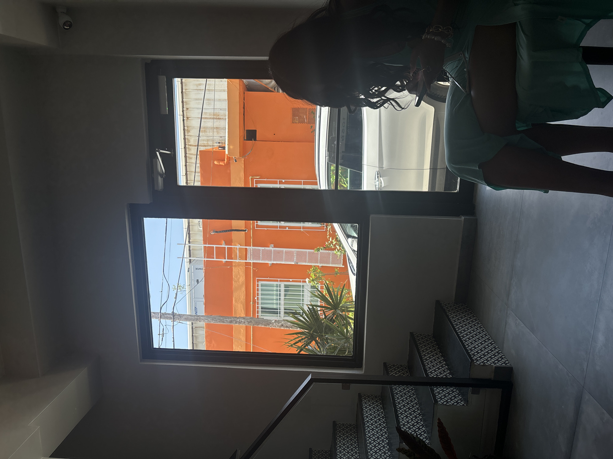 An over-the-shoulder view shows a woman with dark, curly hair sitting in a chair indoors, facing a large window and a glass door. She is silhouetted against the bright outdoor light, holding a smartphone in her right hand. She wears a light teal dress. To her left, a staircase with black and white patterned risers ascends. Through the window and door, an orange building is visible across the street, with a white ladder attached to its side. A white car is parked in front of the building. The floor inside is tiled in large grey squares. The scene is likely in San Juan, Puerto Rico, given the architectural style and vibrant colors common in the city.