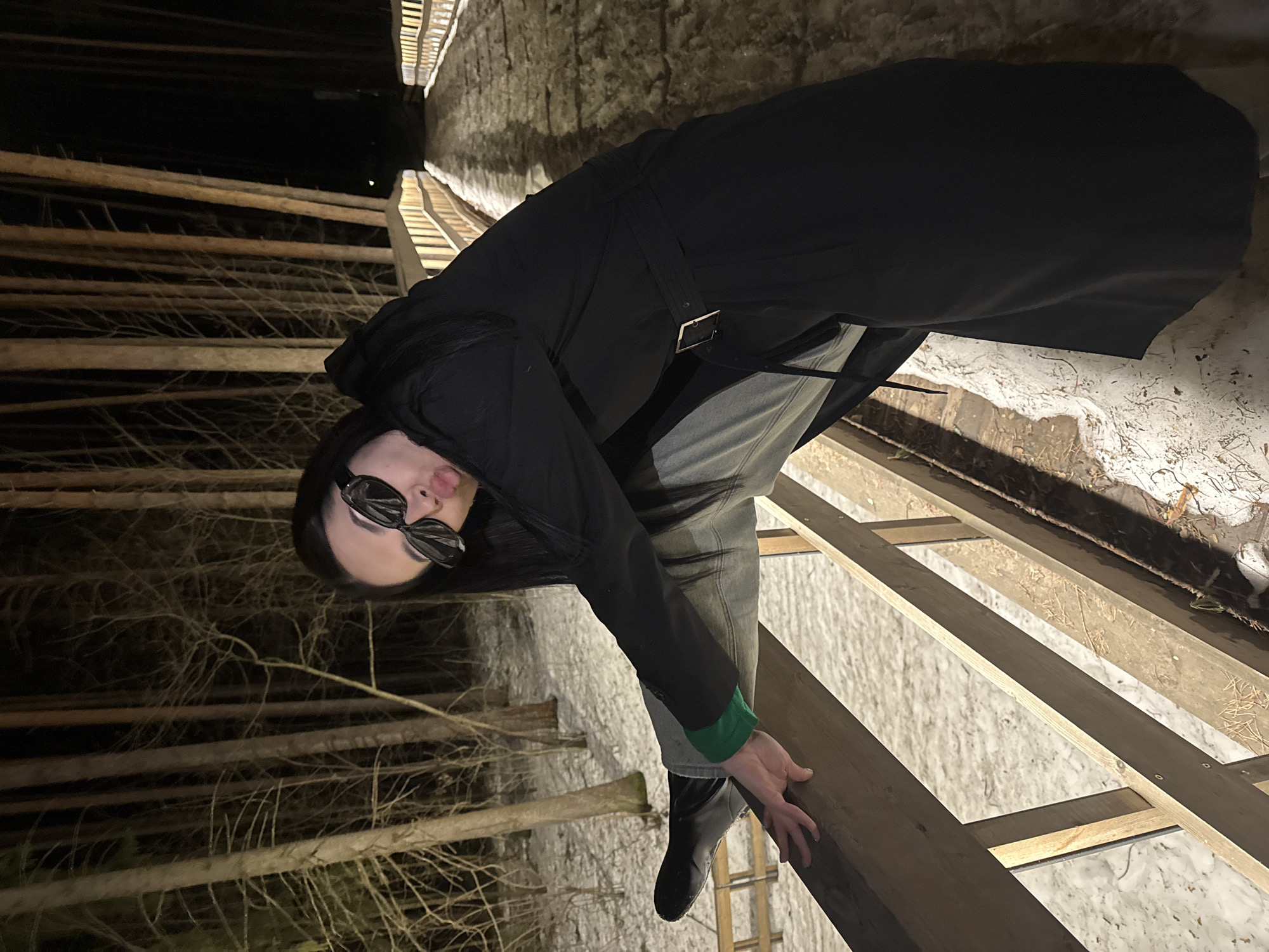 A person with long dark hair and sunglasses, wearing a long black coat with a belt, jeans, and black boots with a green cuff, is standing on a wooden structure. The structure appears to be unfinished, revealing open spaces between the wooden beams. The background consists of trees and snow. The location is identified as Votkinsk, Russia.