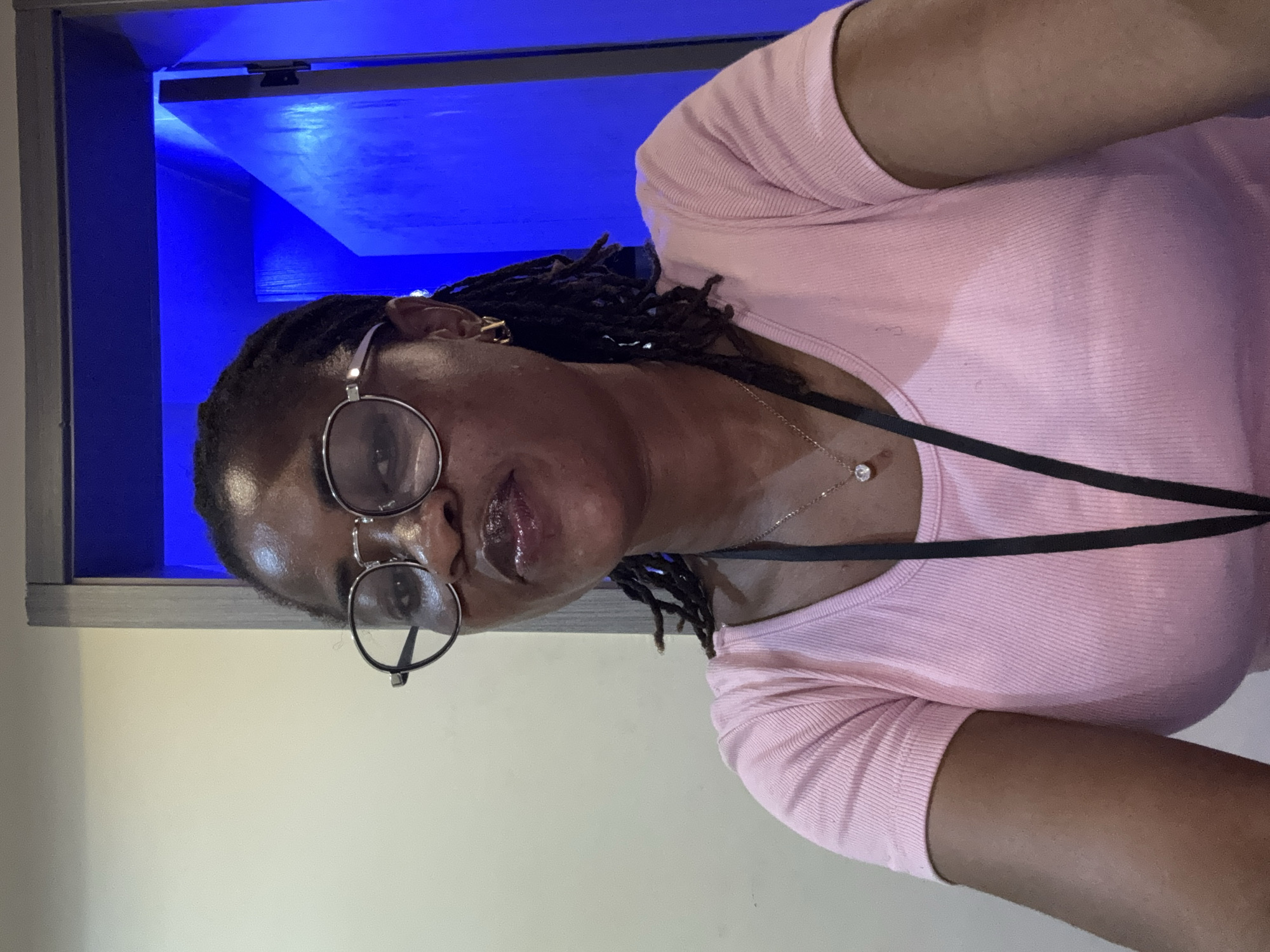 This is a vertically oriented, close-up selfie of a Black woman with locs, wearing glasses and a pink, ribbed t-shirt. She is wearing a delicate necklace with a single clear pendant. Her lips are glossy. She appears to be indoors, in front of a brightly lit blue background. The lighting suggests it might be nighttime or an indoor setting with artificial light. The overall mood is casual and relaxed. The location context provided is Ajara, Nigeria, but there are no specific visual cues in the image to confirm this. There is no visible text in the image.