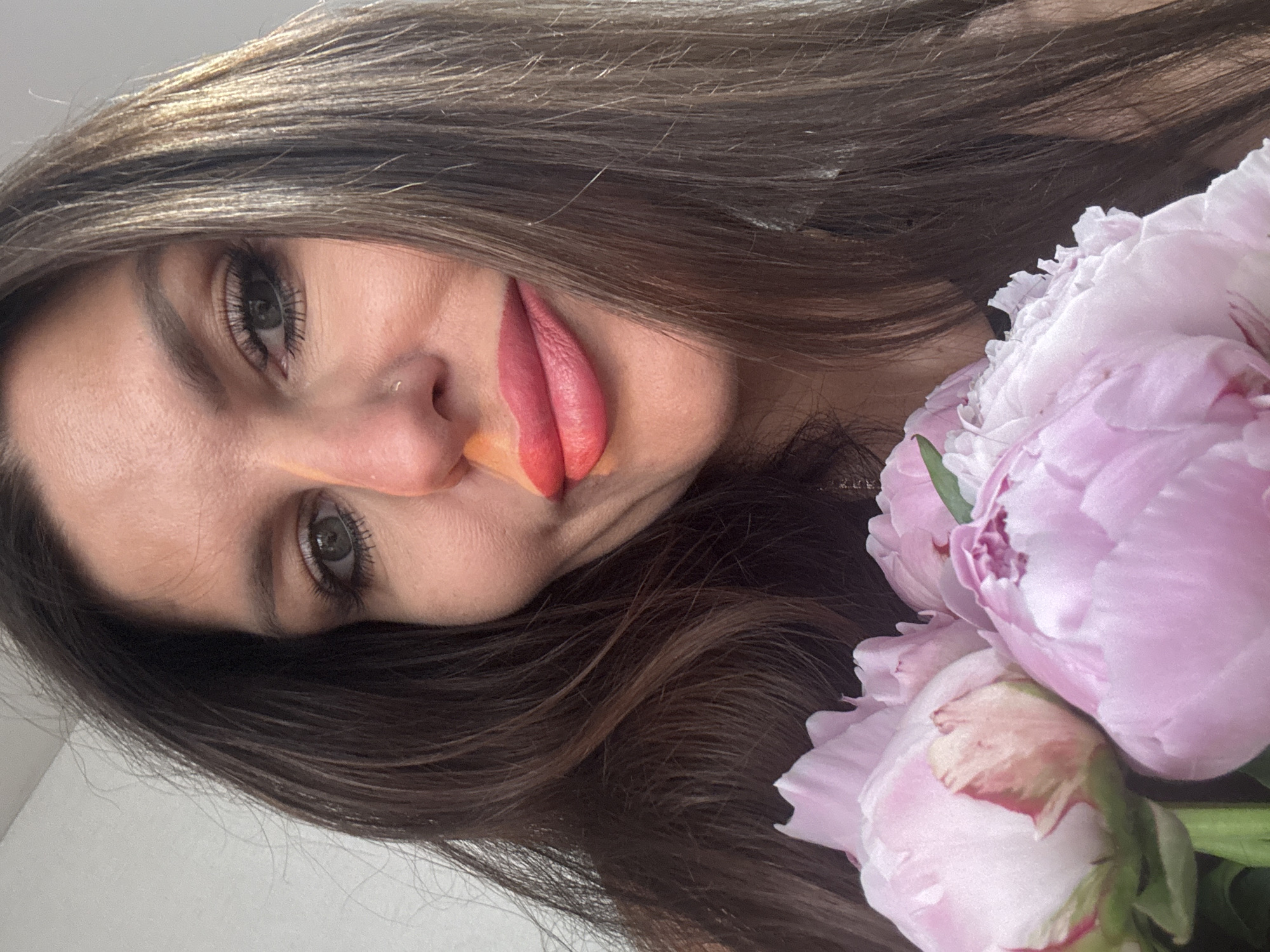 The image is a close-up selfie of a woman with long, dark brown hair, holding a bouquet of pink peonies. She is wearing makeup, including eyeshadow and pink lipstick. Her expression is neutral, and she is looking towards the camera. The background is a plain, light-colored wall. The image does not provide any information about the location in the city of Chaikovsky, Russia.