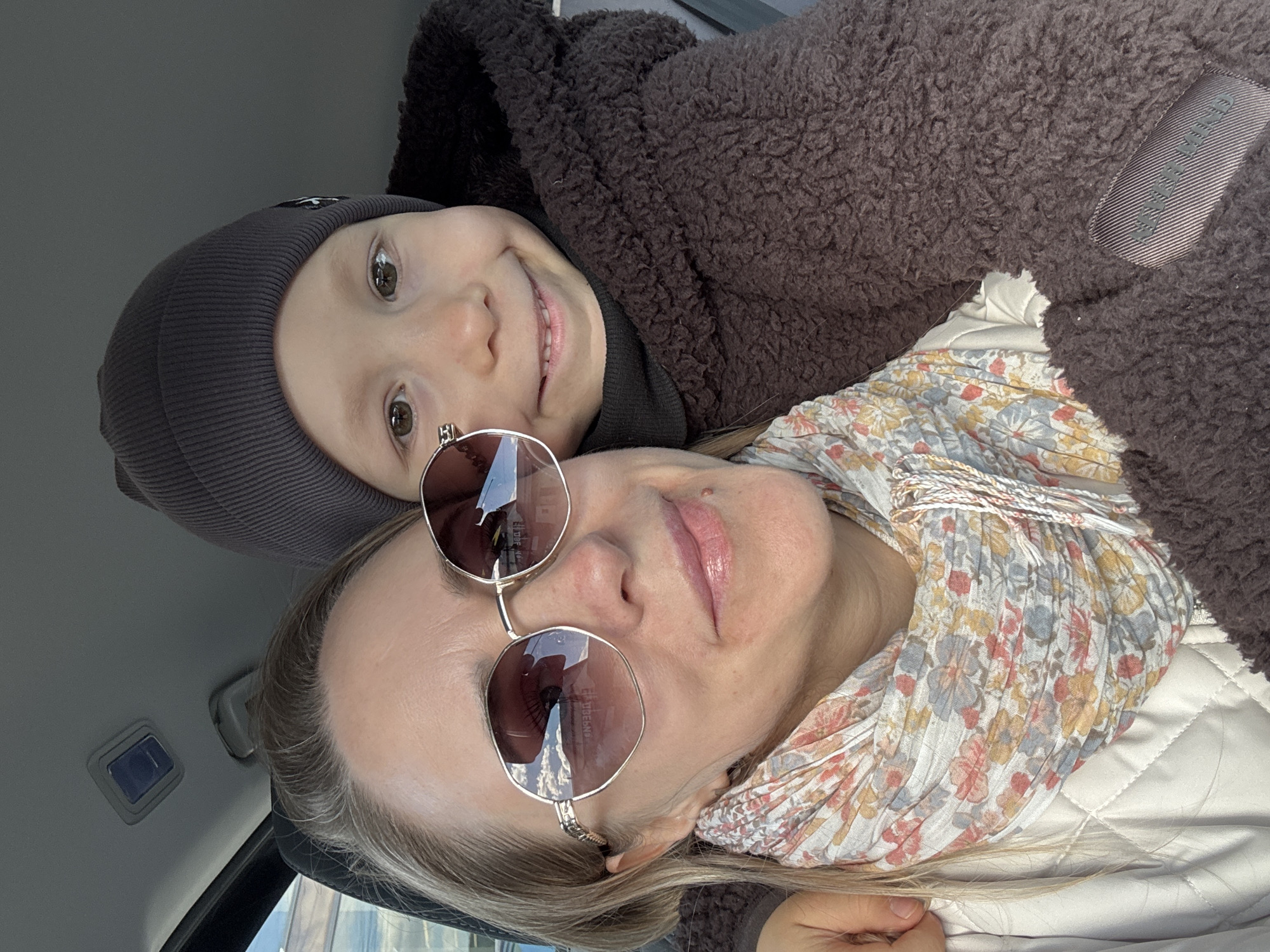 A woman and a child are lying in what appears to be the back seat of a car. The woman is wearing sunglasses and a floral scarf. The child is wearing a dark beanie and is nestled under a brown, fuzzy jacket. The interior of the car is visible, including a light-colored headliner. The location is specified as Chaikovsky, Russia.