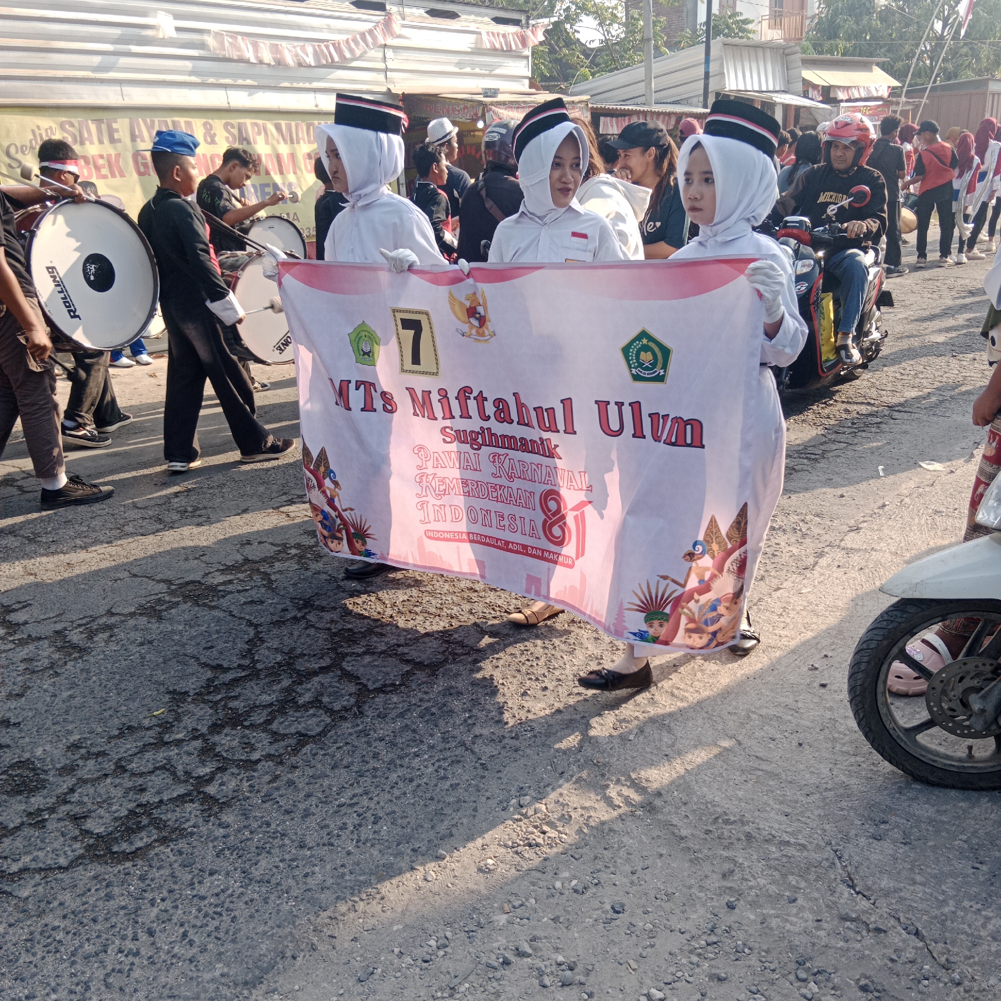 In this medium-shot image, a procession is underway on a cracked asphalt road in Tegalrejo, Indonesia. The sun casts long shadows from the right side of the frame, illuminating the scene. In the foreground, two young individuals, likely students, are holding a large white banner that reads "MTs Miftahul Ulum Sugihmanik Pawai Karnaval Kemerdekaan Indonesia 81" and "INDONESIA BERDAULAT, ADIL, DAN MAKMUR". They are wearing white uniforms with white headscarves and black and red hats, suggesting a religious or educational institution. The banner is adorned with graphics of traditional Indonesian puppets and figures.
Behind them, several individuals are participating in what appears to be a marching band. Some are carrying large bass drums, with one drum displaying the "ROLLING" logo. The participants are dressed in various uniforms, including black and white outfits with similar hats to the banner holders, and some in more casual attire. A person on a red and black motorcycle wearing a red helmet is also part of the procession, moving from right to left. In the background, a banner with Indonesian text, possibly advertising food, is visible, and more people are gathered, observing the event. The overall atmosphere suggests a festive or celebratory occasion, likely a parade for Indonesian Independence Day, given the banner's text. The environment depicts a typical street scene in a town or city.
