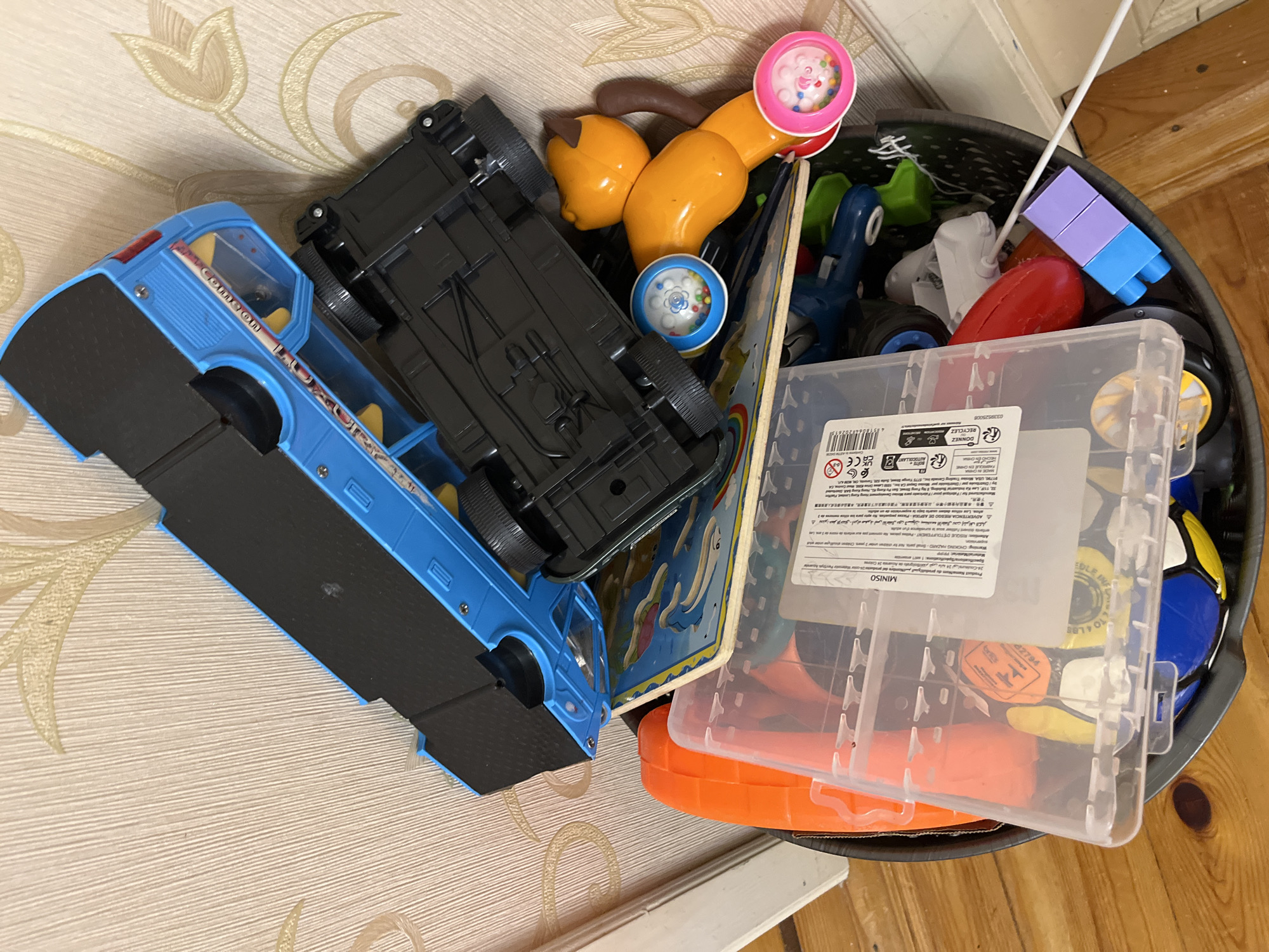 A dimly lit, cluttered toy bin is positioned in a room. The bin, made of dark material, overflows with various toys. Prominently featured are two large toy vehicles, one blue and one black, stacked on top of each other. A bright orange toy, possibly a cone, sits to the left of the bin. A clear plastic container with a white lid, filled with smaller toys, rests on top of the bin's contents. A yellow and brown stuffed animal with small, colorful rattles attached to its limbs is also visible. Several other toys, including a blue and yellow ball, a red oval-shaped object, and a small purple and blue block structure, are scattered throughout the bin. The background consists of a cream-colored wall with a faint floral pattern and a wooden floor. The scene appears to be an indoor setting in an unspecified location.