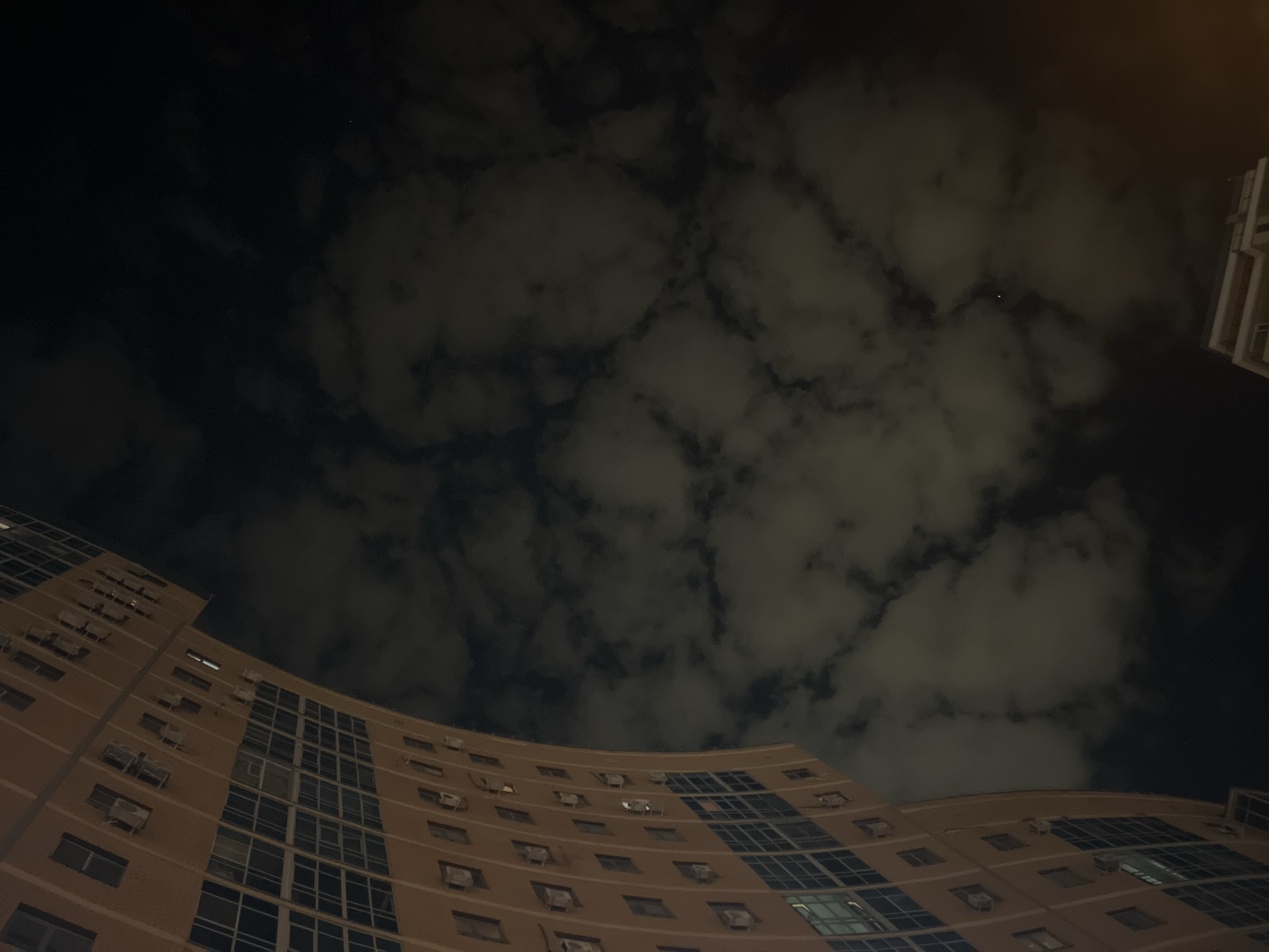 The image shows a low-angle view of a multi-story building against a cloudy night sky. The building appears to be of modern construction with a curved facade. It is illuminated by artificial light. Each window has its own split air-conditioning unit. The sky is mostly dark with clouds present, some with the appearance of a city glow. There is another building peeking out from the top-right. There is a single bright dot above the buildings that can either be a star or an airplane. The location is Kazan, Russia.