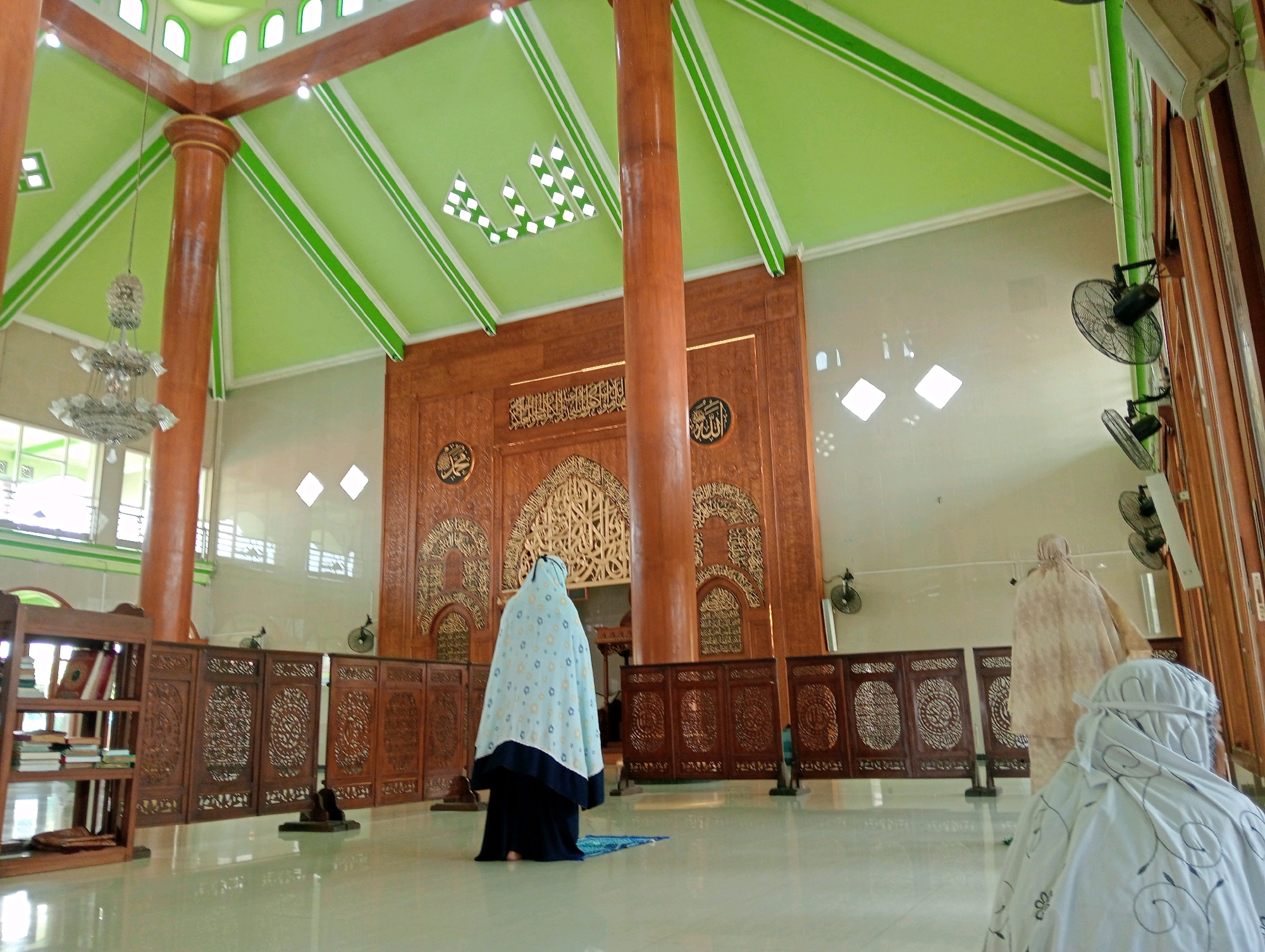 The image shows the interior of a mosque in Soko, Indonesia, with a bright green ceiling and large wooden pillars. Three women are present, all wearing hijabs. One woman, in the foreground and facing away from the camera, is wearing a white hijab with black floral patterns and appears to be praying. To her left, another woman in a light blue and white patterned prayer dress and dark headscarf is also facing away, on a blue prayer mat. Further back and to the right, a third woman in a light brown patterned hijab is walking away from the camera. The mosque features ornate wooden screens and a decorative wooden mihrab. Natural light enters through windows and is supplemented by a chandelier and ceiling lights. Fans are mounted on the walls. To the left, a wooden bookshelf is visible, filled with books.