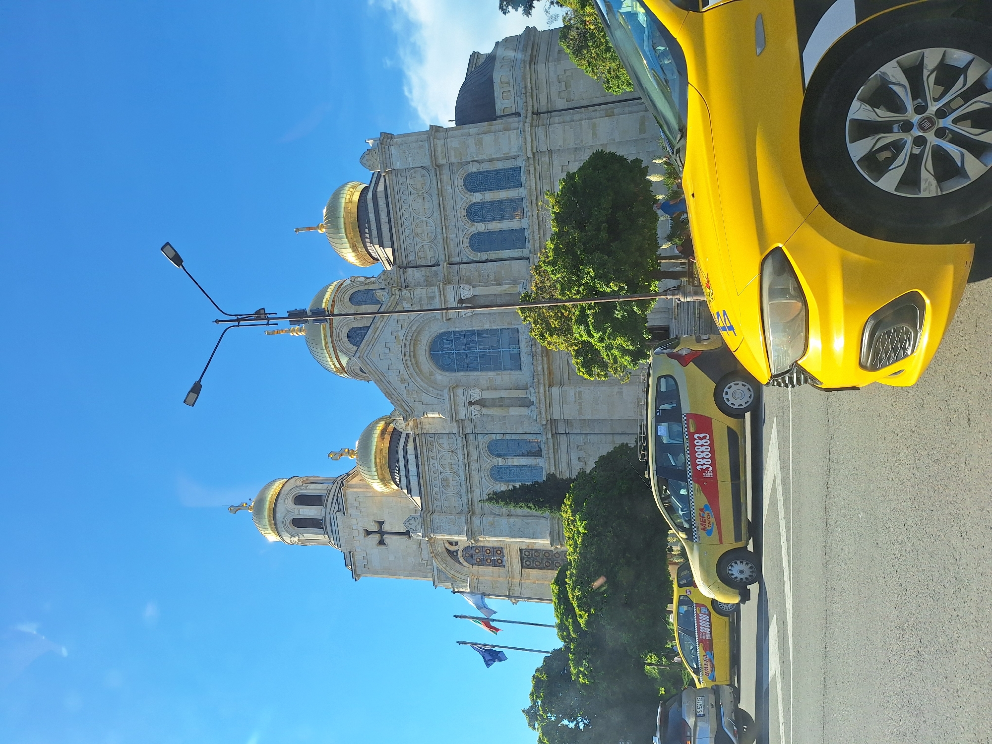 The image depicts the Cathedral of the Assumption of the Virgin in Varna, Bulgaria, on a sunny day. Several yellow taxis are visible on the street in the foreground. The cathedral, a prominent landmark, features golden domes and intricate architectural details. The sky is clear blue with a few wispy clouds. The scene appears to be a typical urban street view with the cathedral serving as a backdrop to daily life, including vehicular traffic.