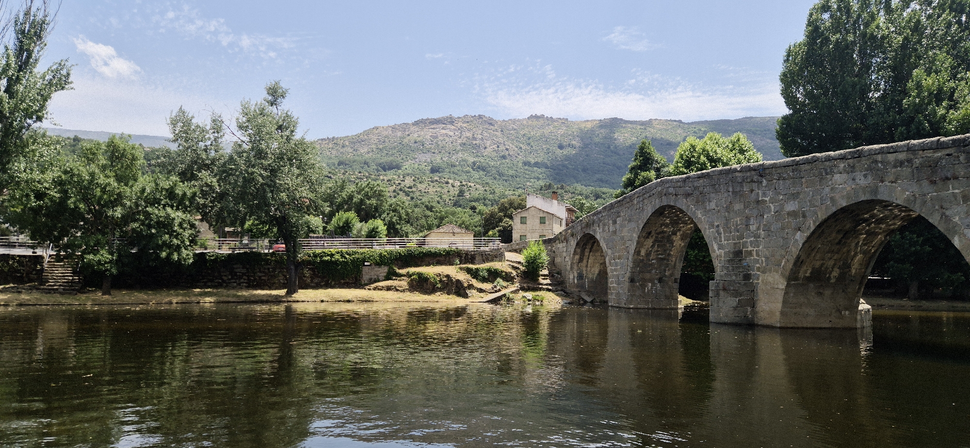 This is a wide-angle, eye-level photograph of an old stone bridge spanning a calm body of water. The bridge, constructed from rough-hewn, grey stones, features multiple arches, with the largest and most prominent one dominating the right side of the frame. Sunlight casts dappled shadows beneath the archways, hinting at a bright day.
The scene is lush and green, with mature trees lining the left bank of the water. Their leaves are a vibrant green, suggesting it is likely springtime or summer. In the background, rolling hills covered in dense vegetation rise towards a hazy, mountainous horizon. A light blue sky with scattered white clouds stretches above, indicating fair weather.
On the left bank, nestled amongst the trees, are a few small buildings. One appears to be a light-colored house with a red-tiled roof and green shutters. A stone retaining wall with some ivy growing on it runs along the edge of the water, and a set of stone steps leads up from the water's edge. A white railing follows the edge of the bank. There are no people visible in the image. The overall atmosphere is peaceful and serene, capturing a tranquil rural landscape. The time of day appears to be midday, given the bright sunlight.