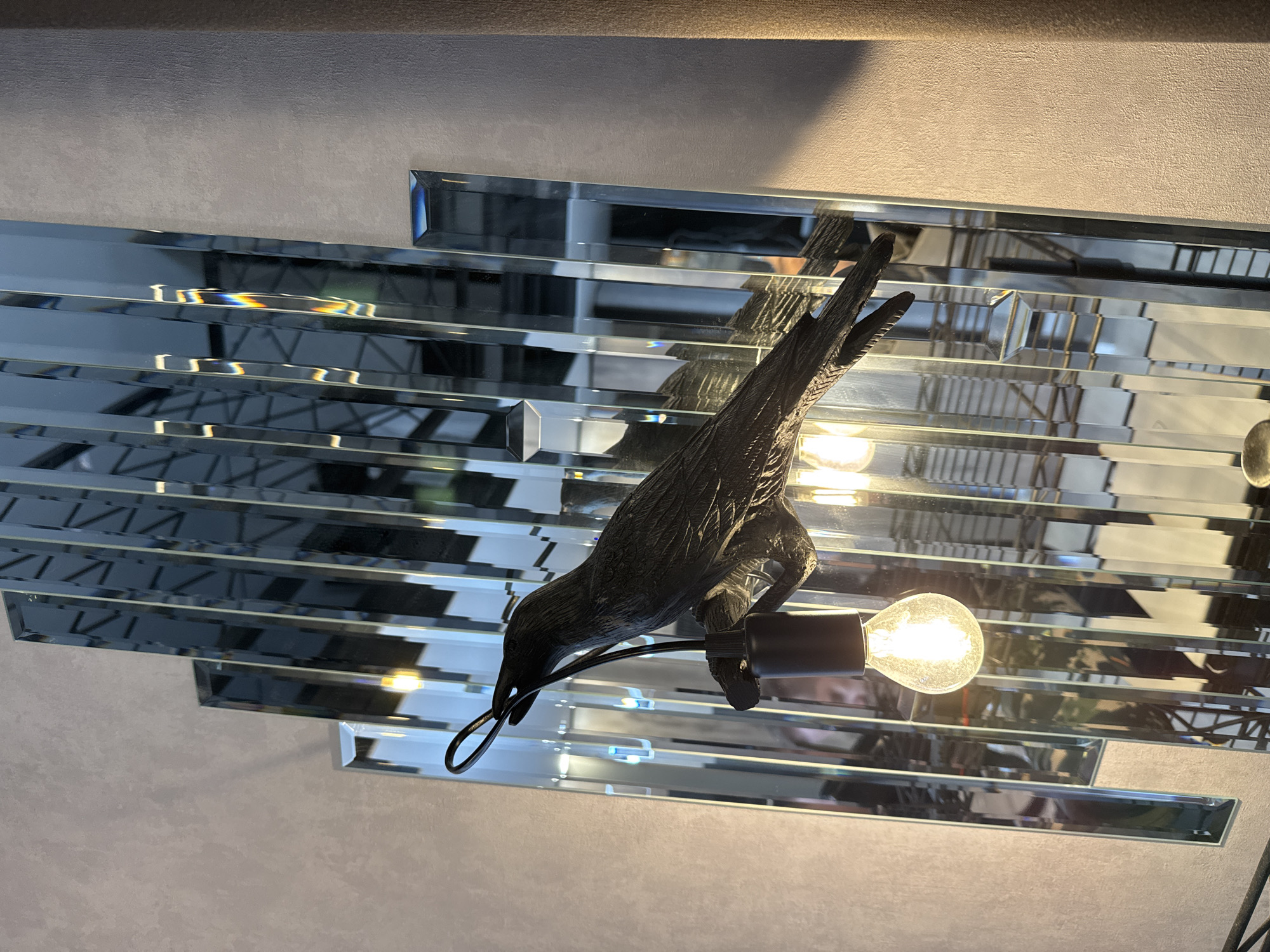 This image presents a low-angle view of a distinctive ceiling or wall-mounted light fixture. The central element is a dark, intricately sculpted bird, resembling a crow or raven, which appears to be clutching an illuminated incandescent light bulb in its beak. The bird exhibits textured details, suggesting materials like carved wood or dark metal.
The background is composed of multiple rectangular mirrored tiles arranged in horizontal, slightly offset rows, creating a layered and reflective surface. These tiles capture and refract light, producing various reflections and geometric patterns, including a subtle rainbow-like effect in some areas. Above and to the sides of the mirrored section, a plain, textured surface, likely a wall or ceiling painted in a light neutral tone, is visible.
A second, partially visible light bulb on the right indicates that the bird and its bulb are part of a larger lighting installation. The scene is indoors, showcasing a decorative lighting piece that contributes to a modern or eclectic interior design aesthetic. No human figures or discernible activities are depicted. The reliance on artificial light makes the time of day indeterminate, and there is no visible text.