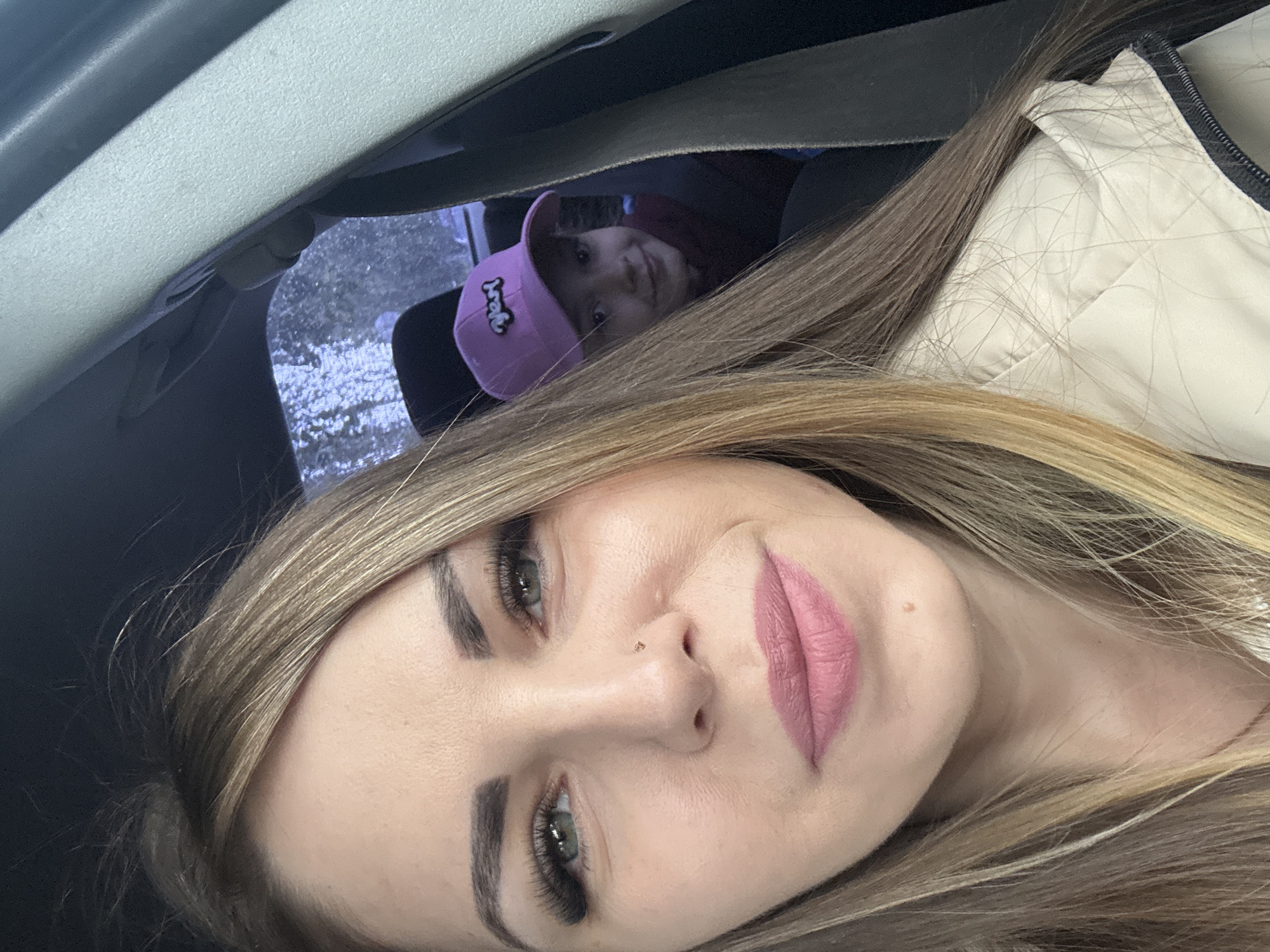 A selfie photograph taken from inside a vehicle in Razdolye, Russia. The primary subject is a woman with light brown hair, fair skin, and pink lipstick. She has green eyes and a nose piercing. Behind her, visible between the woman and the roof of the car, is a child wearing a pink baseball cap. Outside the car window is snow-covered foliage. The interior of the car, including the seat belt, roof, and headrest, are visible. The woman is wearing a light beige quilted jacket.