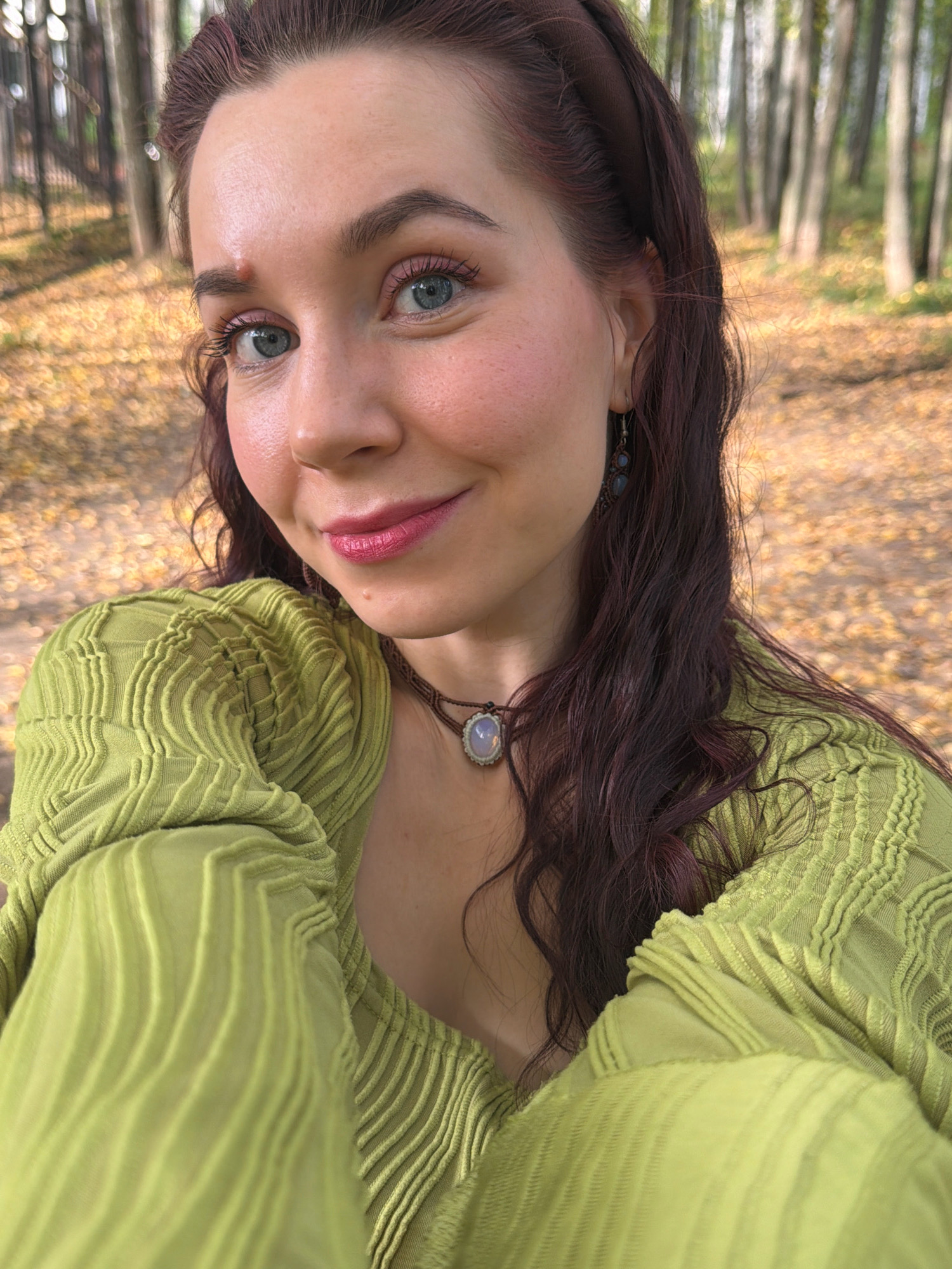 A medium shot selfie features a young woman with auburn hair and blue eyes. She is smiling and looking directly at the camera. She is wearing a light green, textured blouse and a necklace with a light-colored pendant. Her makeup includes pink blush and lipstick. The background is blurred, showing trees and fallen leaves, suggesting an outdoor setting in an autumnal environment, possibly a park. The lighting is natural and appears to be daytime. The image does not depict any specific identifiable landmarks or scenes from the city of Chaikovsky, Russia.