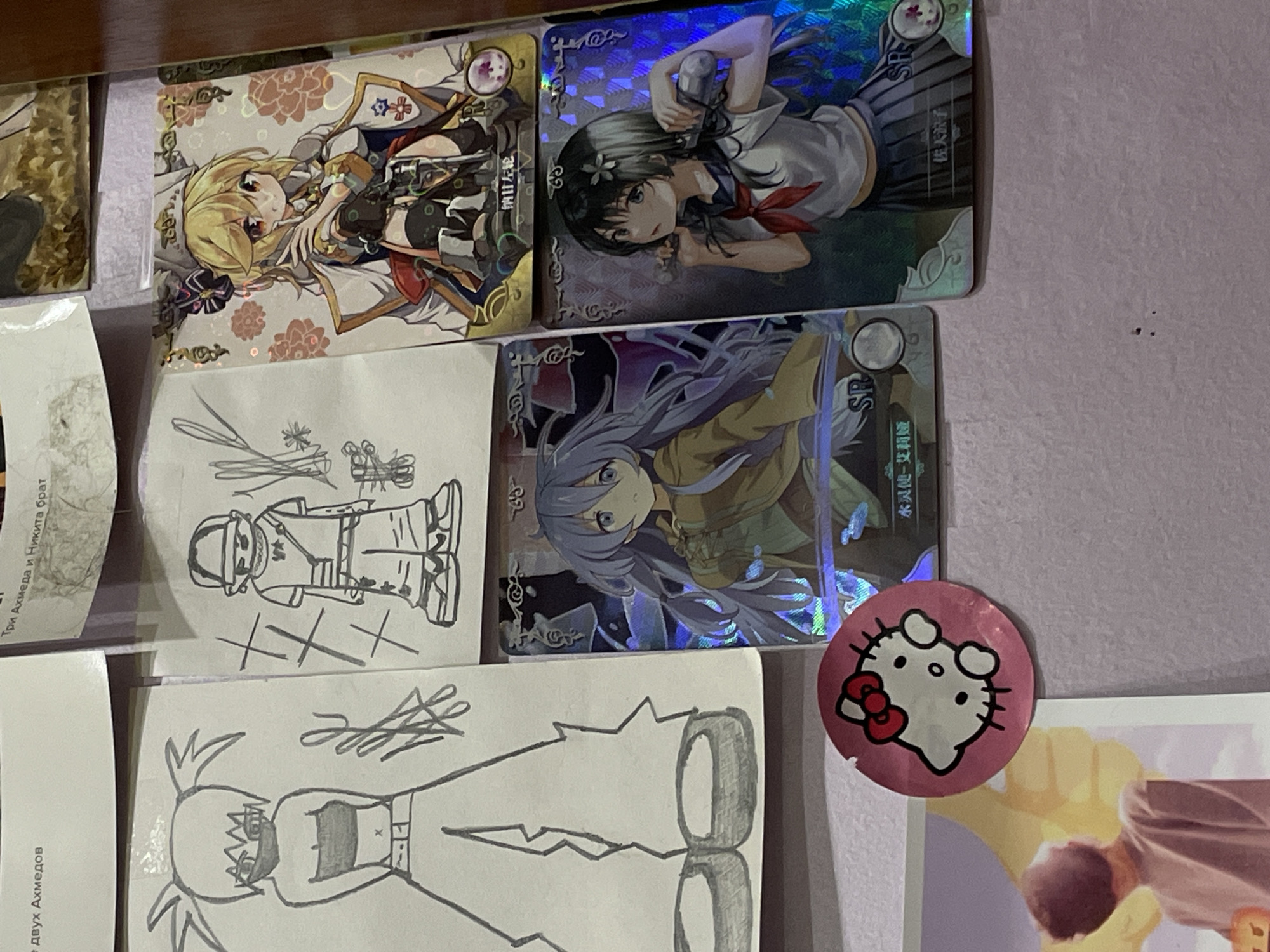 The image displays a collection of anime-style trading cards and drawings arranged on a flat surface, likely a desk or table. In the upper left corner, text in Russian is partially visible. To the left of the trading cards, two hand-drawn sketches of anime characters are present. The trading cards themselves feature colorful illustrations of female anime characters. A pink Hello Kitty sticker is adhered to the surface in the lower left portion of the image. In the lower left of the image, a portion of a poster or print is visible, showing the back of a person looking upwards. The overall scene appears to be a personal collection of artwork and merchandise. There is no discernible indication of a specific location within a city, including ЖК «Борисоглебское», Russia, or any specific urban context. The image focuses solely on the displayed items.