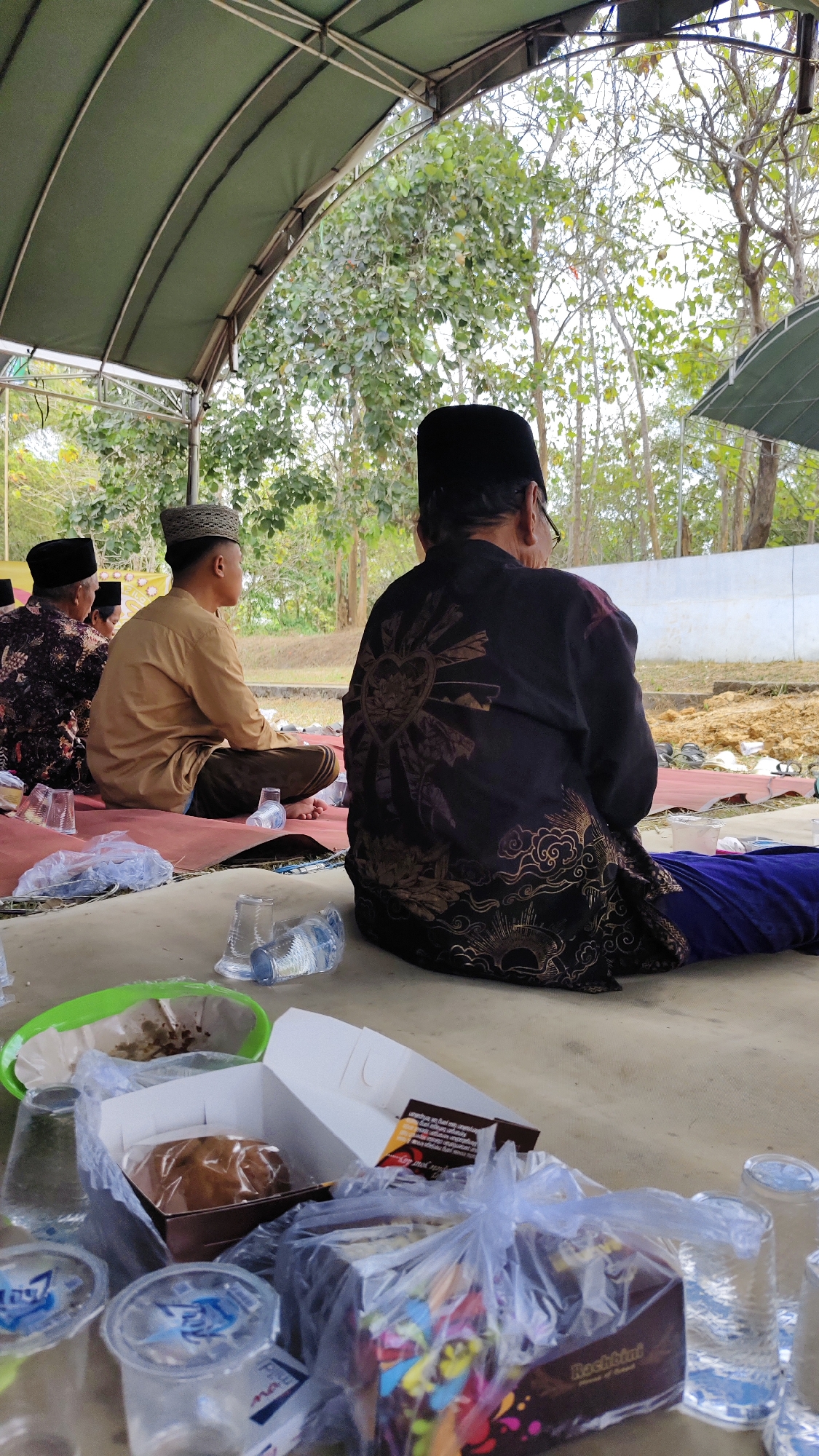 A medium shot captures an outdoor gathering under a green canopy. Three men are seated on a red mat on the ground, facing away from the camera. The man in the foreground, on the right, wears a black peci (a traditional cap) and a black shirt adorned with a gold sunburst design with a lotus flower in the center. His head is tilted down, and he is wearing glasses. To his left, a younger man in a tan shirt and a patterned peci sits with his back to the camera, looking to the side. Further left, another man, partially visible, wears a black patterned shirt and a peci. The ground is covered with a light-colored mat, and scattered around are plastic cups, some containing water, and white take-out boxes with food. In the background, trees with sparse foliage are visible against a bright sky, and a white wall is present. The overall setting suggests a communal meal or event in a semi-rural area, possibly in Telagah, Indonesia, given the traditional attire of the men.